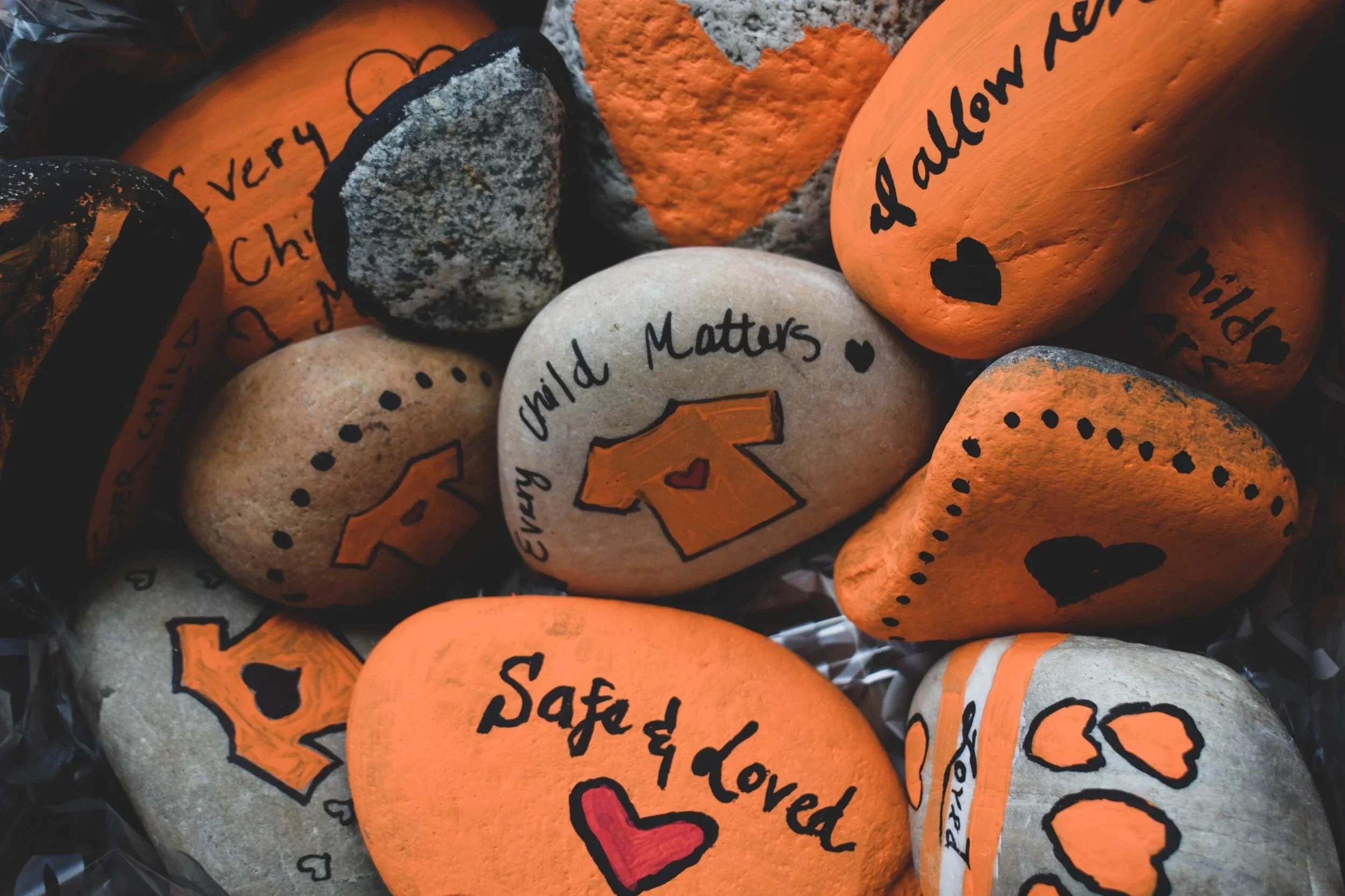 Multiple painted rocks with mixed colors of orange, white, and black. The rocks feature handwritten messages and designs, including hearts and words like 'child', 'love', 'safety', and 'kind'.