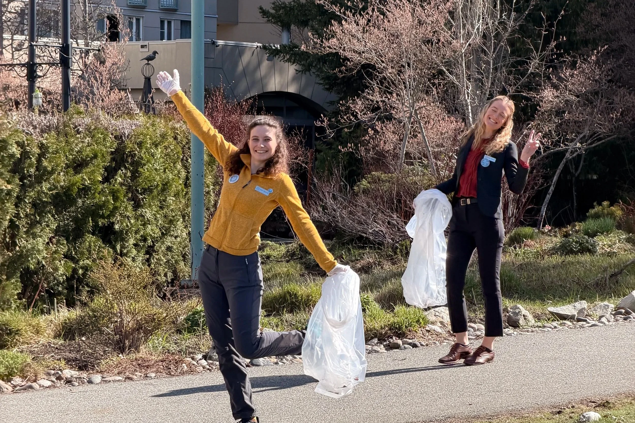 Two women outdoors, one wearing a yellow jacket and the other in a black suit, participating in a community cleanup event, holding trash bags.