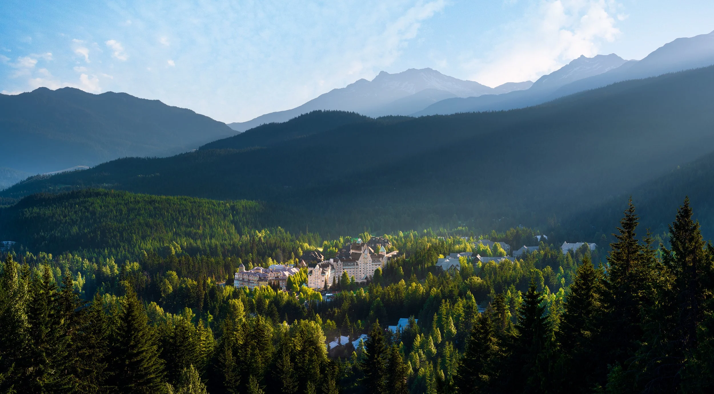 Scenic view of Fairmont Chateau Whistler in the heart of the coast mountains nestled in a forested valley beneath a partly cloudy sky.