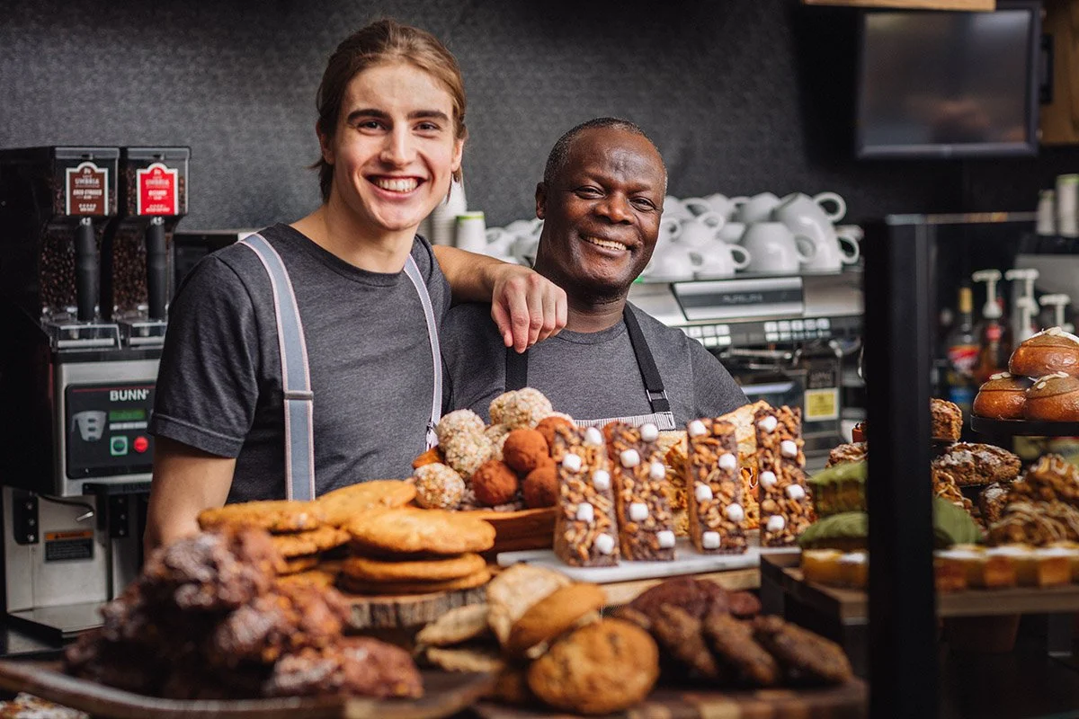 Two smiling men standing behind a display of various baked goods, including cookies, bars, and truffles, inside Portobello coffee shop