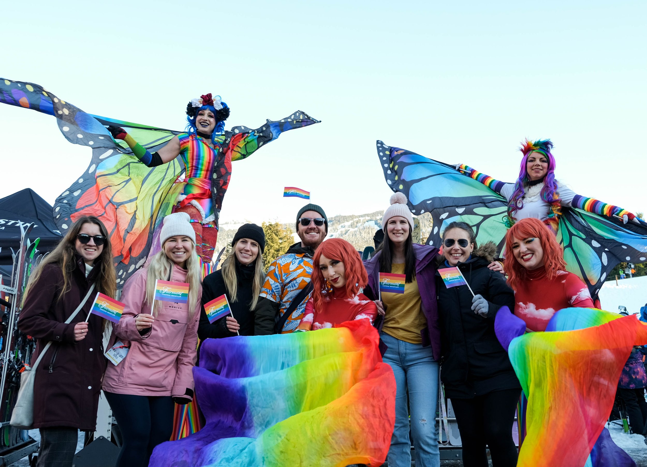 Group of people at a pride parade, with two individuals dressed as colorful butterfly fairies with rainbow wings standing on stilts, surrounded by others holding rainbow flags and rainbow streamers, outdoors in winter.