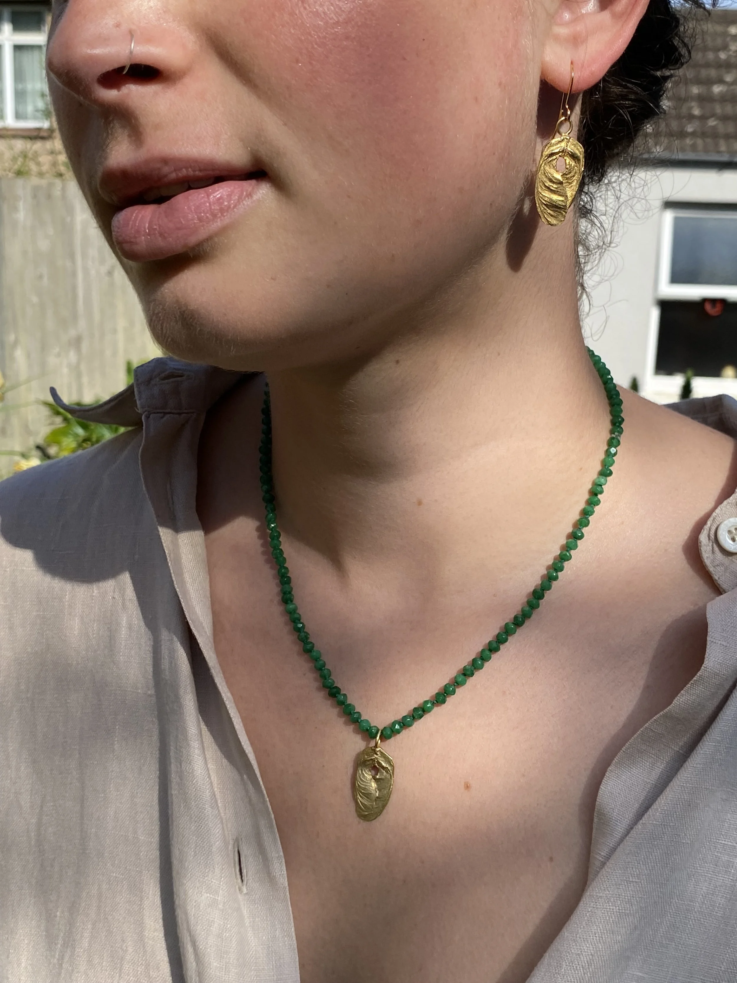 Close-up photo of a woman wearing a gold earring and a matching gold necklace with a green beaded chain, outdoors in sunlight.