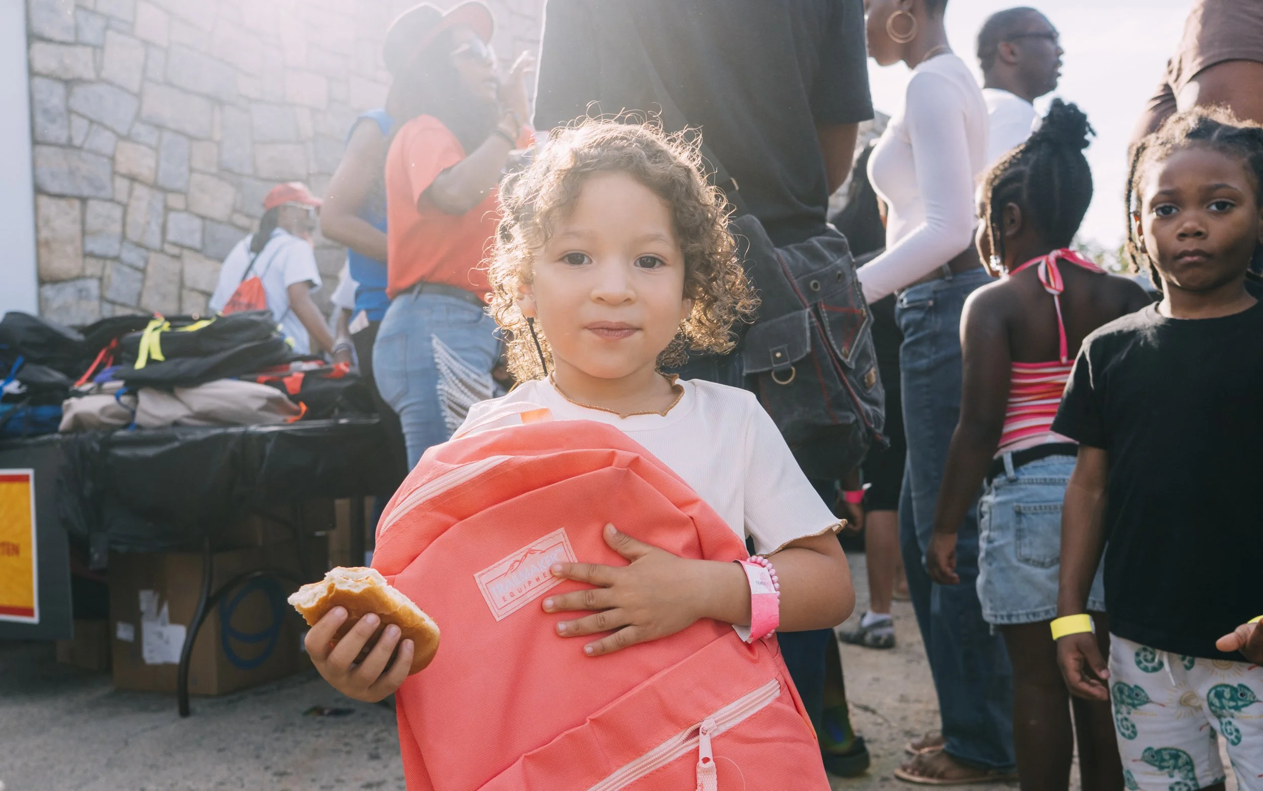 Young girl holding a pink backpack and eating a sandwich, standing in front of a group of people near a table with backpacks and supplies, outdoors during daytime at Naeem Justice Event