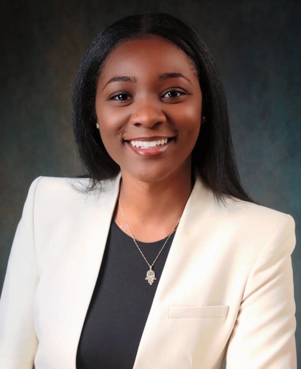 Professional portrait of a young African American woman smiling, wearing a white blazer, black top, and a silver necklace with a Hamsa charm, against a dark background.