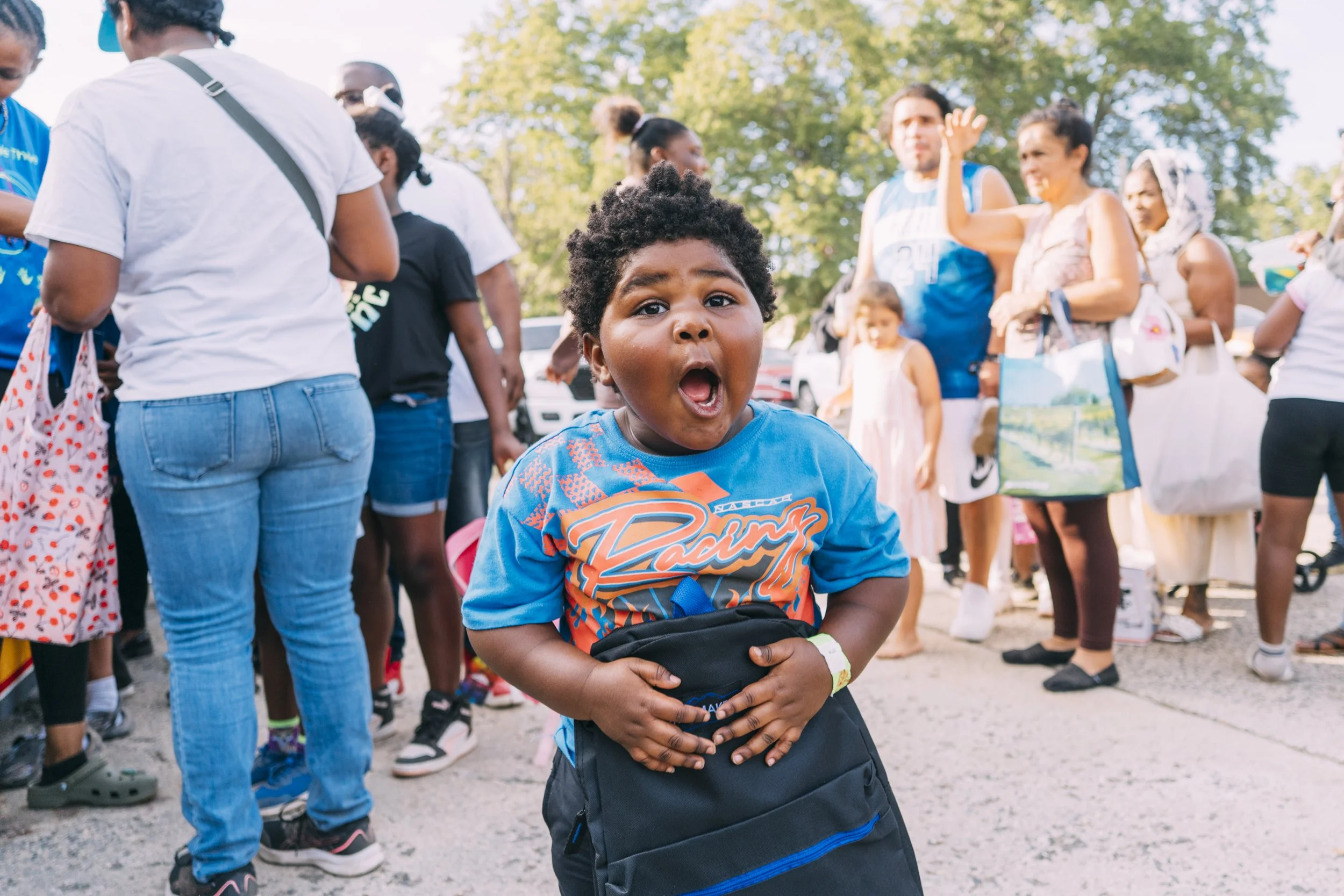 A young boy with curly hair and a surprised expression, holding a backpack in front of a crowd of people outdoors during daytime.