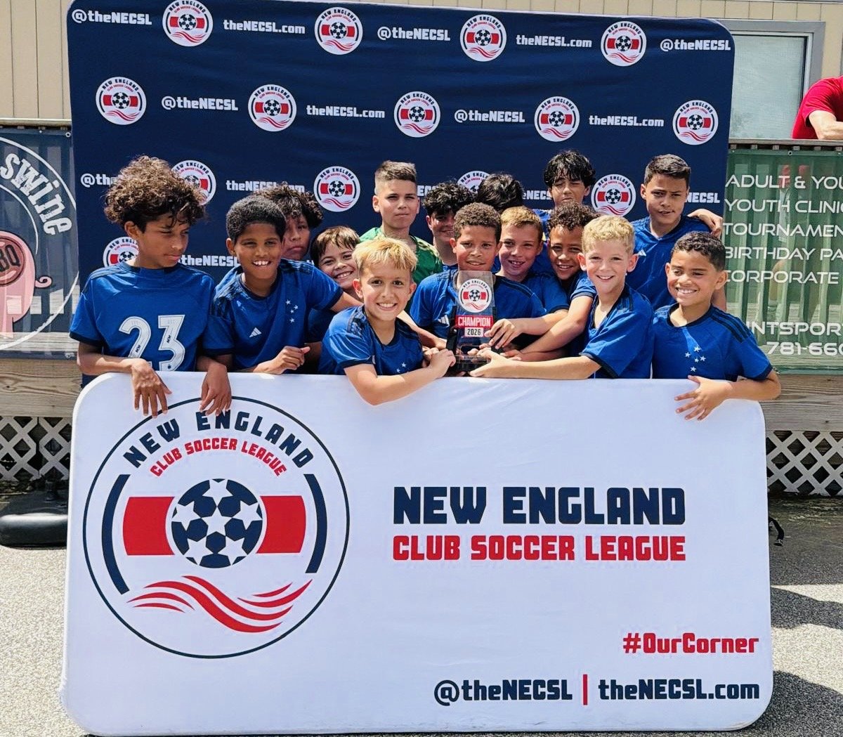Young boys in blue soccer uniforms holding a trophy and standing behind a large sign that reads 'New England Club Soccer League' and features a soccer ball logo. The background shows a blue banner with the league's social media handle and website.