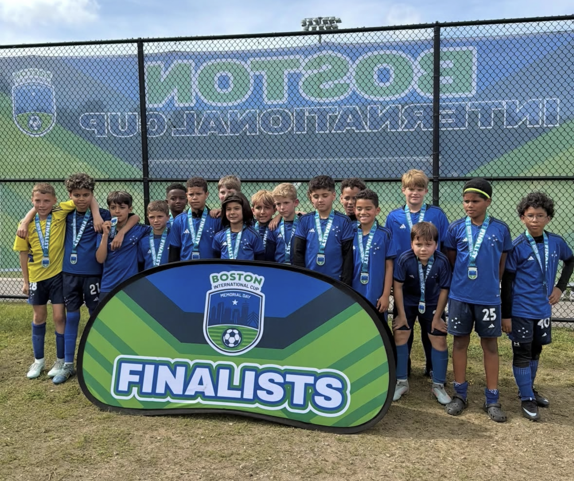 Youth soccer team standing on a field wearing blue uniforms and medals, posing behind a sign that reads 'FINALISTS' with a green and blue design, against a chain-link fence with a sponsor banner in the background.