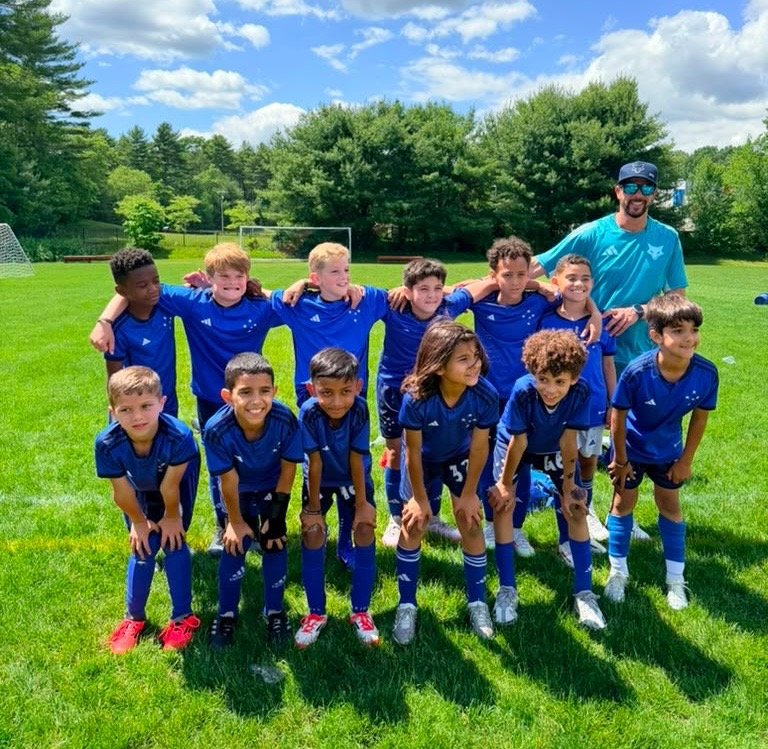 A youth soccer team in blue jerseys and shorts posing on a soccer field with their coach. The team has ten children, standing and kneeling, under a blue sky with white clouds, surrounded by green trees.