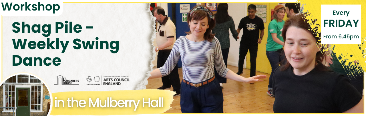 Room of people practicing swing dance, with two women at the forefront. One wearing a white and black striped t-shirt, with blue jeans and a brown belt. The other is in a black t-shirt. Both have brown hair.