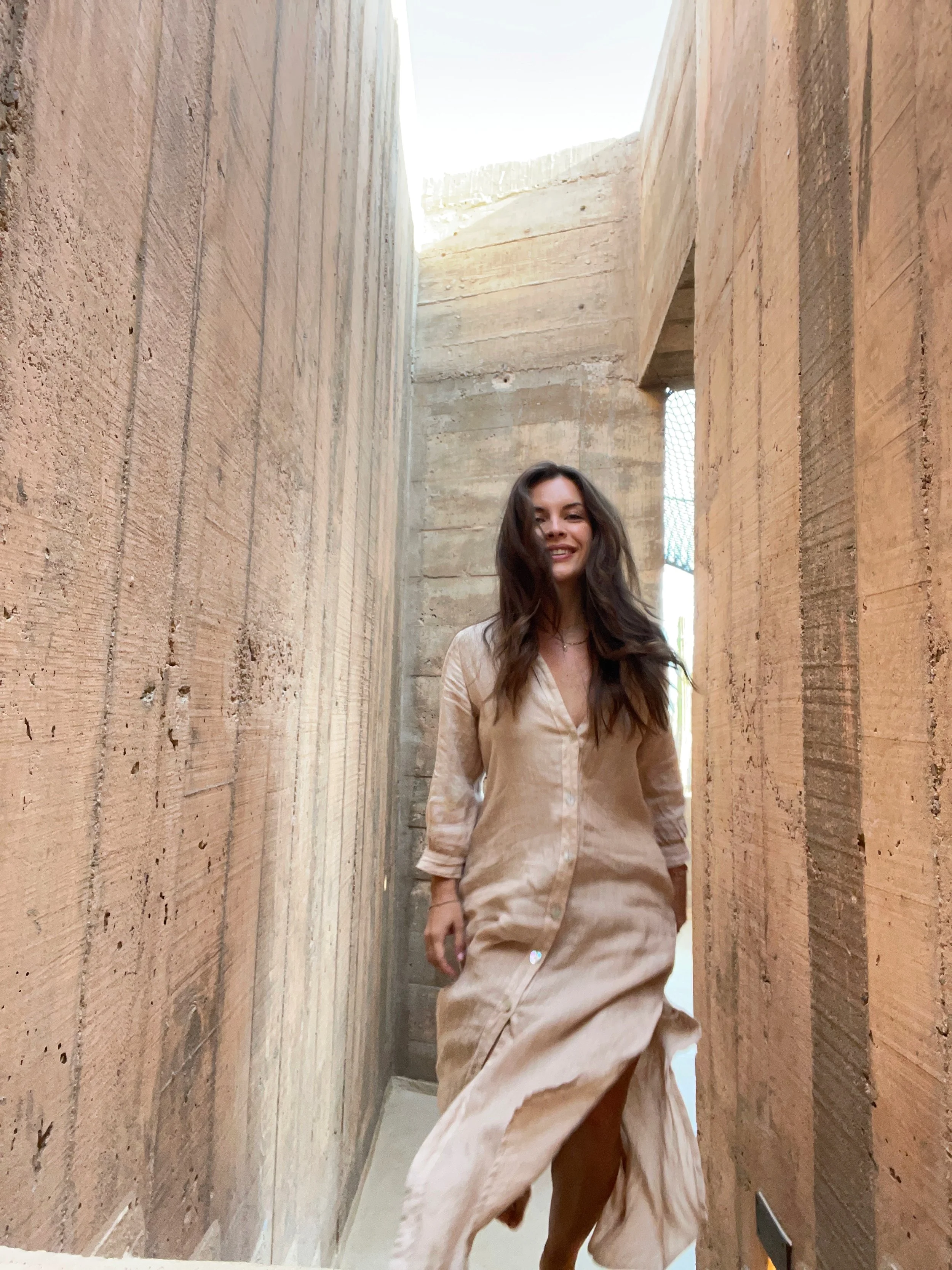 A woman with long brown hair smiling while walking through a narrow space with tall, textured beige walls.