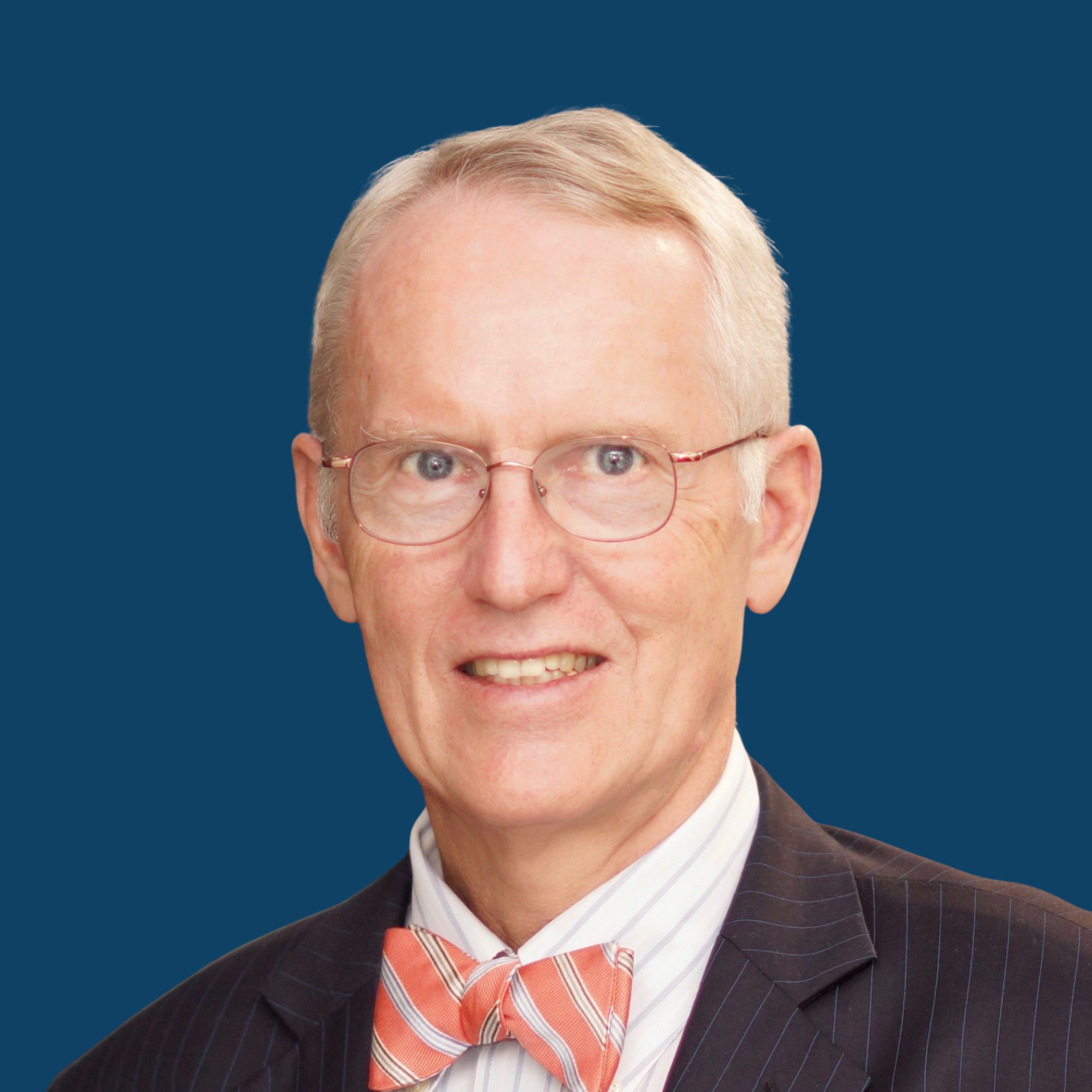 Portrait of an elderly man with glasses, short gray hair, dressed in a pinstripe suit, white shirt, and a pink and white striped bow tie, against a plain blue background.