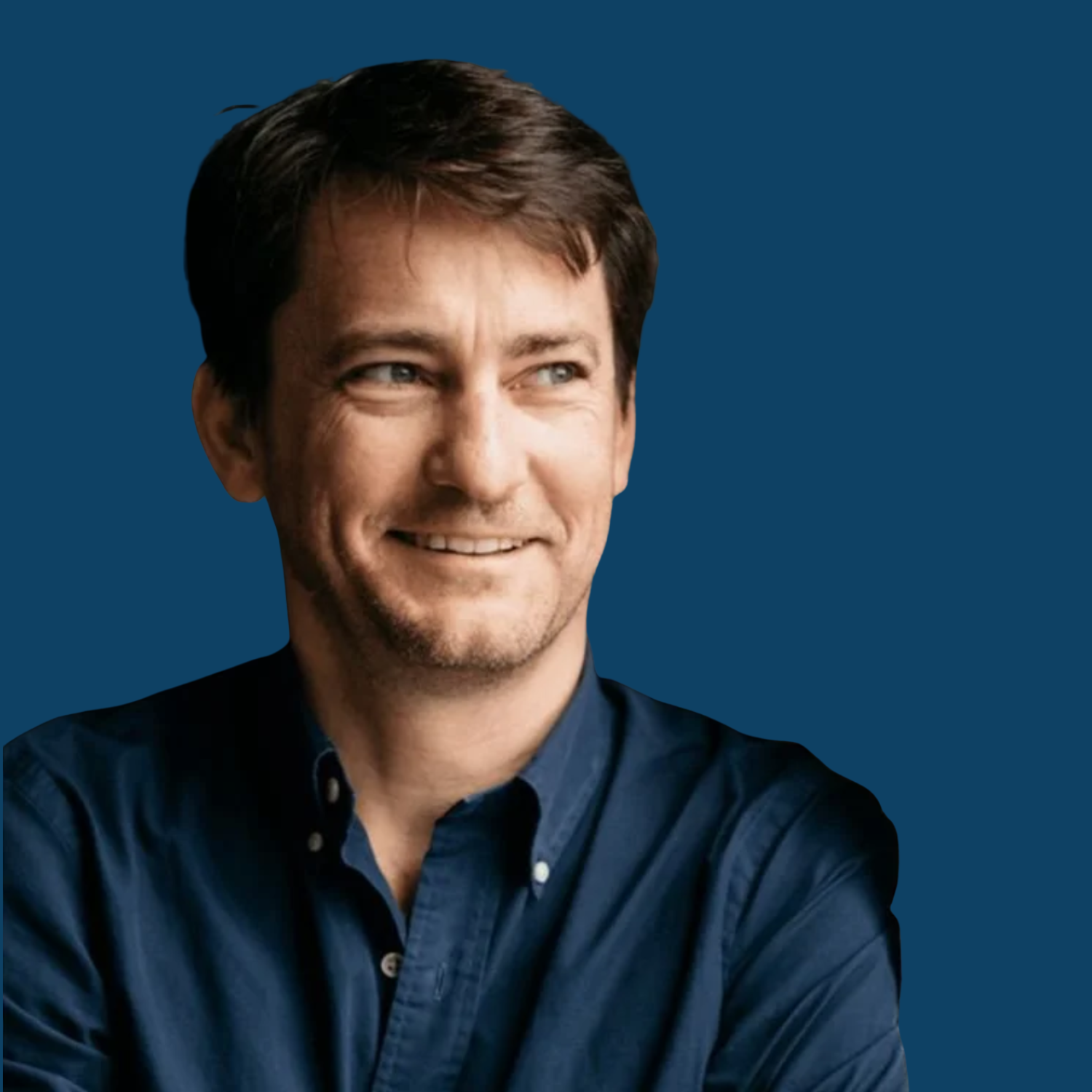 A man with dark brown hair and blue eyes smiling, wearing a dark blue shirt against a solid blue background.