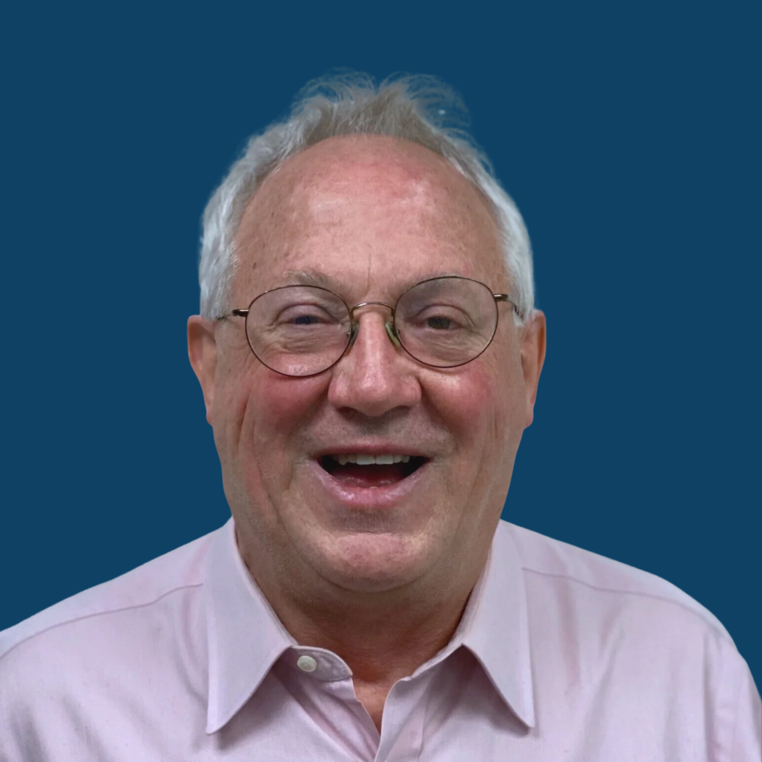 A smiling older man with glasses and grey hair, wearing a light pink collared shirt, against a solid dark blue background.