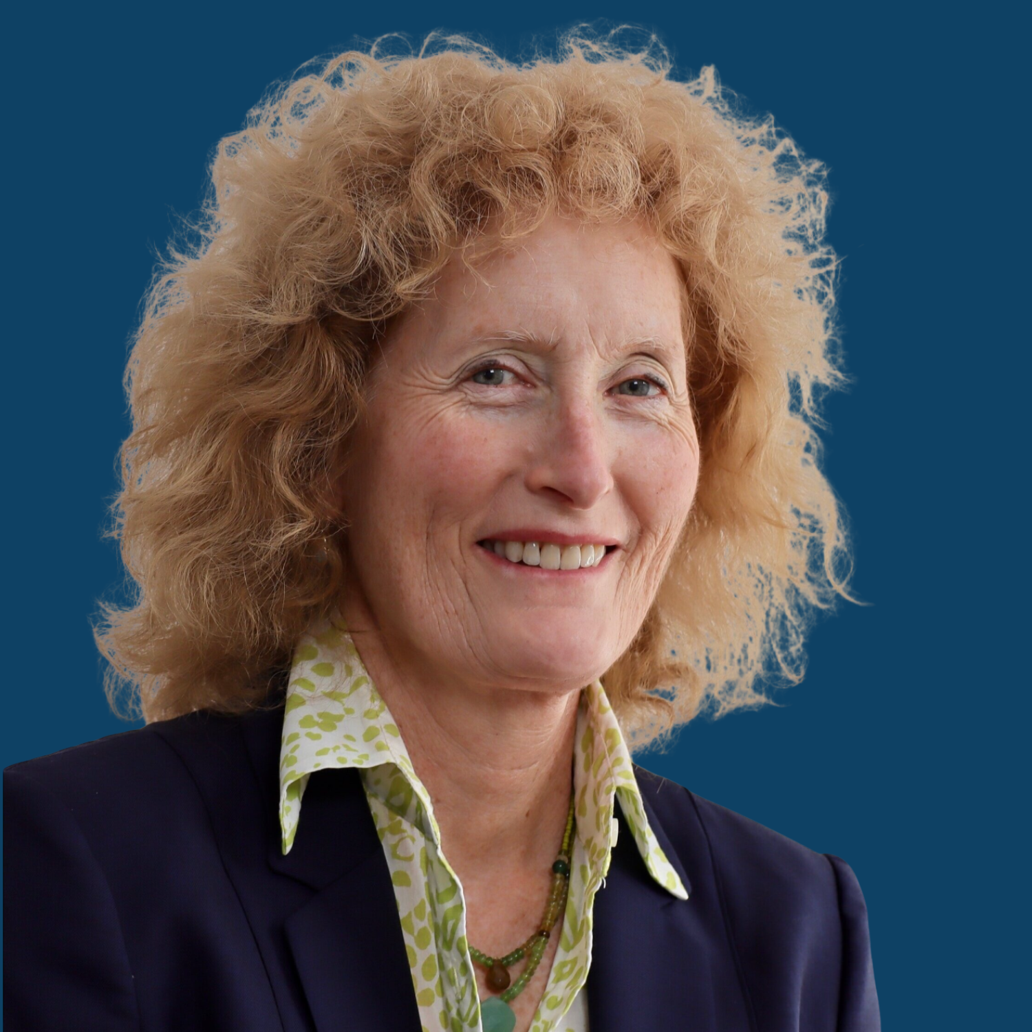 A woman with curly blonde hair smiling, wearing a dark blazer over a white and green patterned shirt, and a beaded necklace, against a solid blue background.