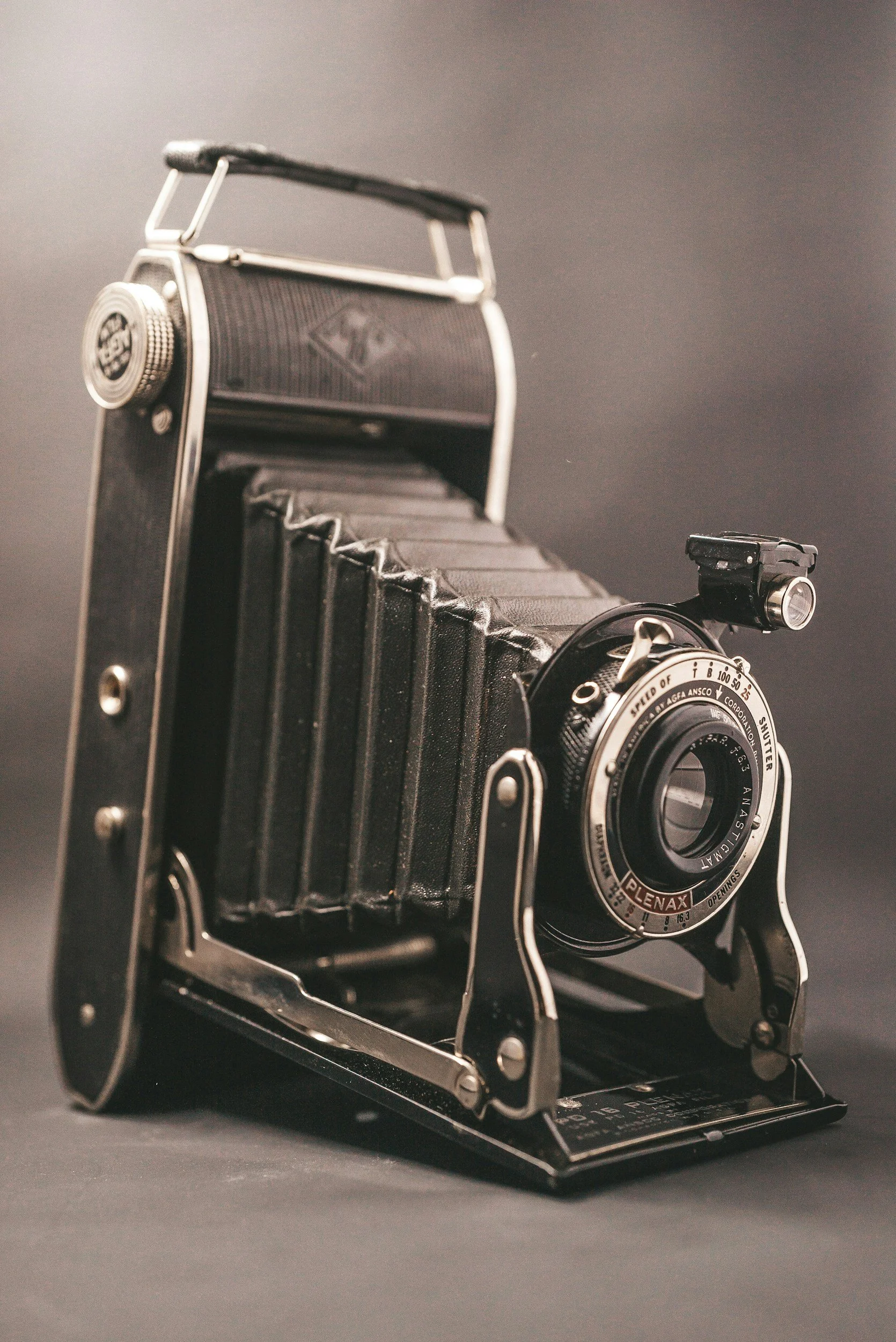 A vintage film camera with a black body, silver accents, and a bellows design, placed on a black surface against a gray background.