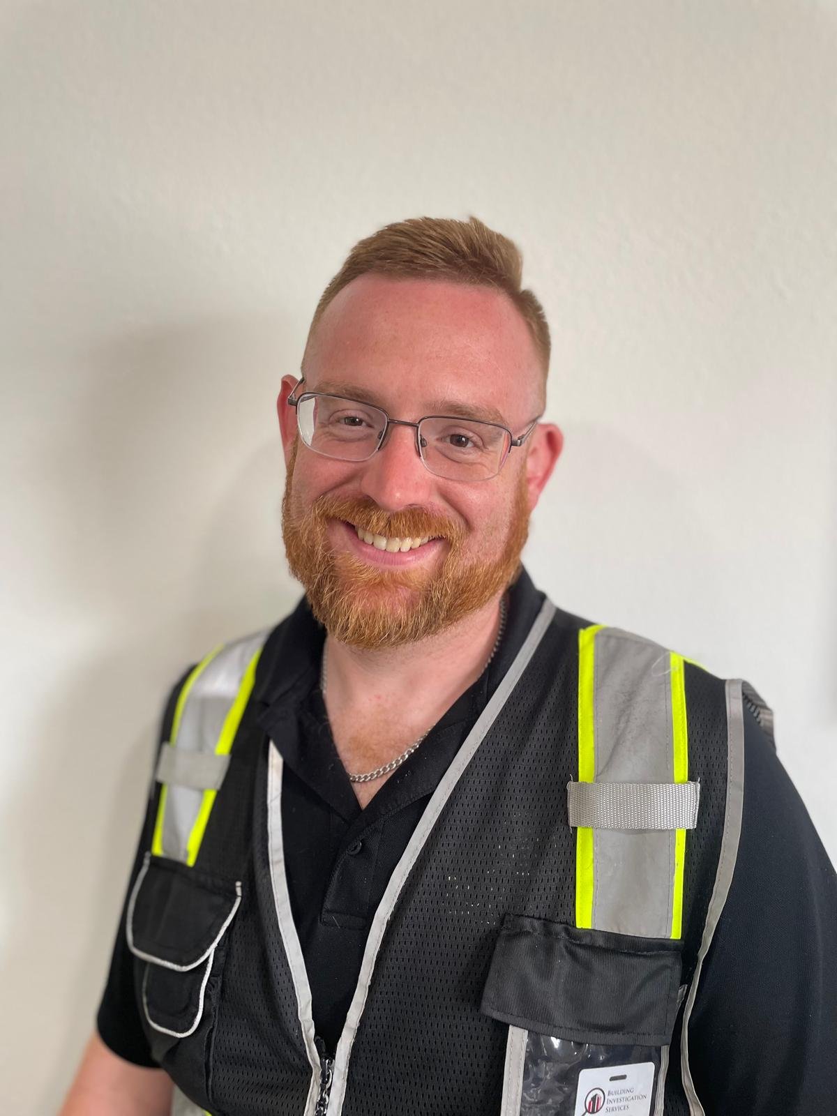 A smiling man with red hair, beard, and glasses wearing a black uniform and a reflective safety vest, standing against a plain white wall.