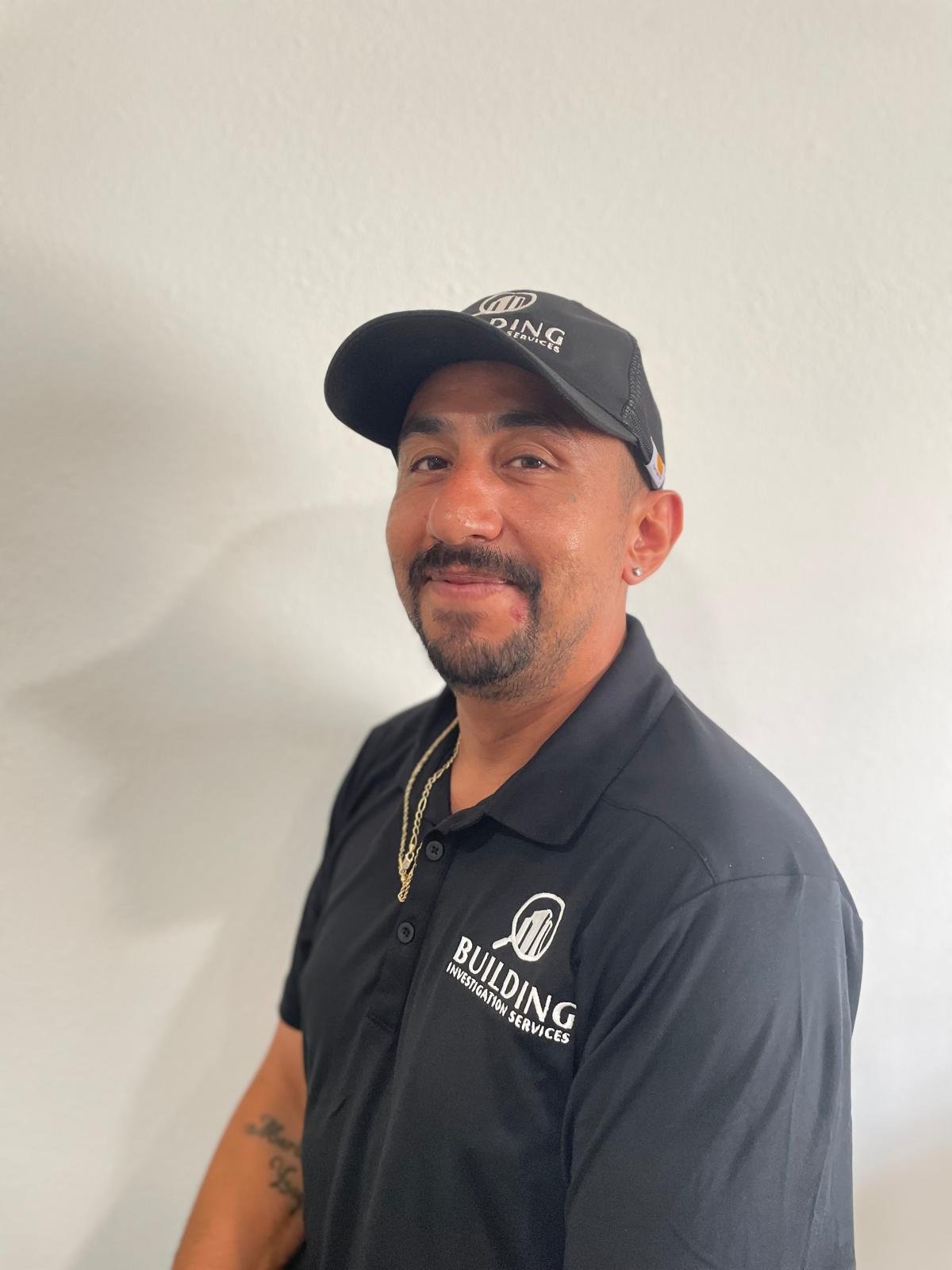 A man with a mustache, beard, and earrings wearing a black cap and black polo shirt with building investigation services logo posing against a plain wall.
