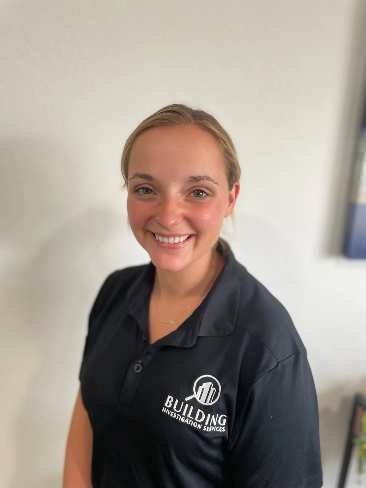 A young woman with blonde hair smiling, wearing a black polo shirt with a logo for Building Investigation Services.