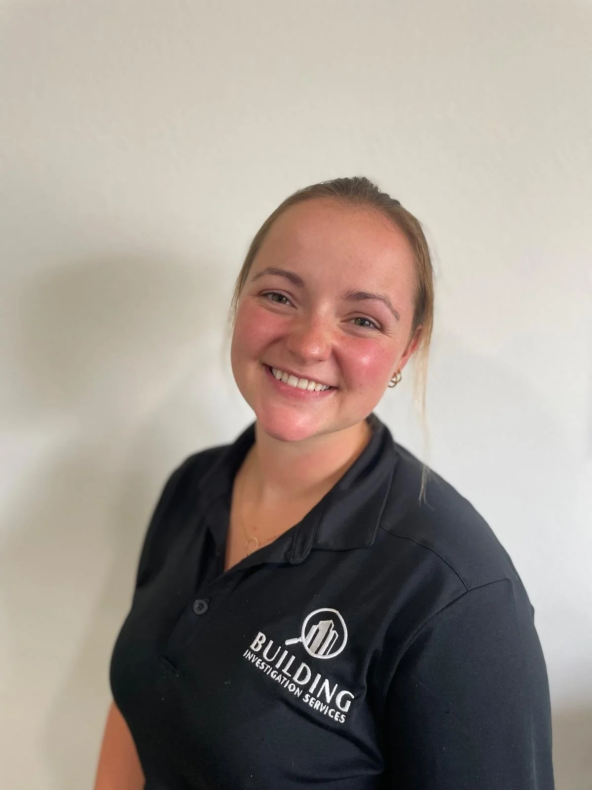 A smiling young woman with light skin, wearing a black polo shirt with the logo and text 'BUILDING Investigation Services' on it, standing against a plain white wall.