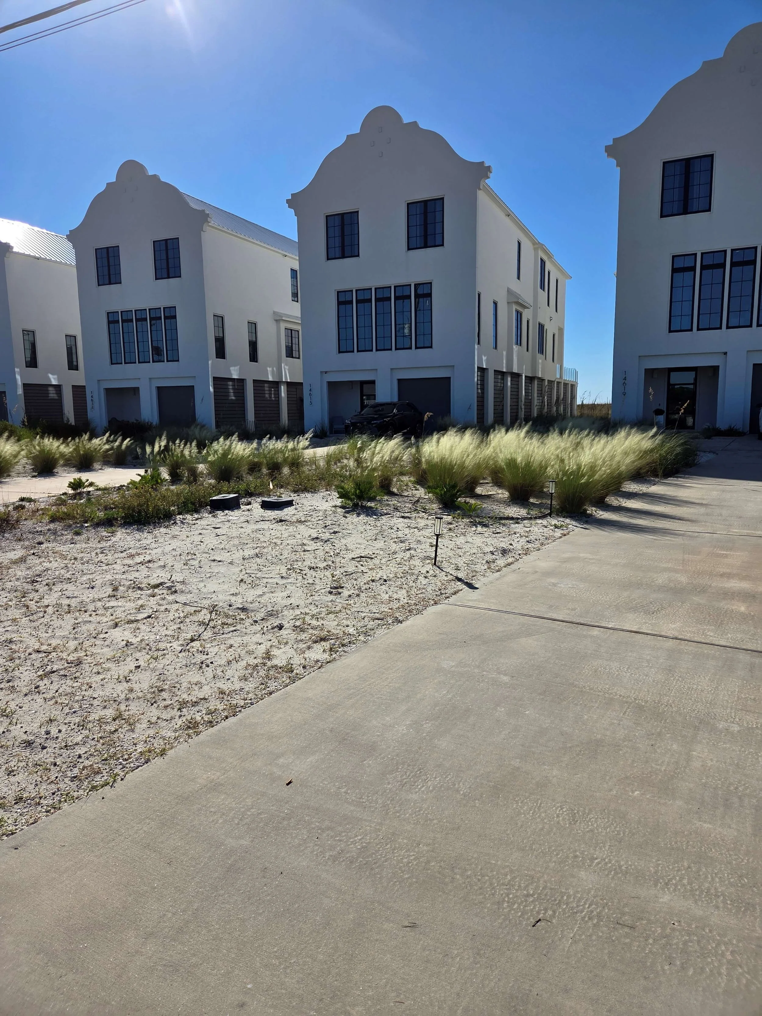 White modern homes with black window frames and garage doors, sandy landscaping with grasses, a black vehicle, and a concrete sidewalk under a clear blue sky.