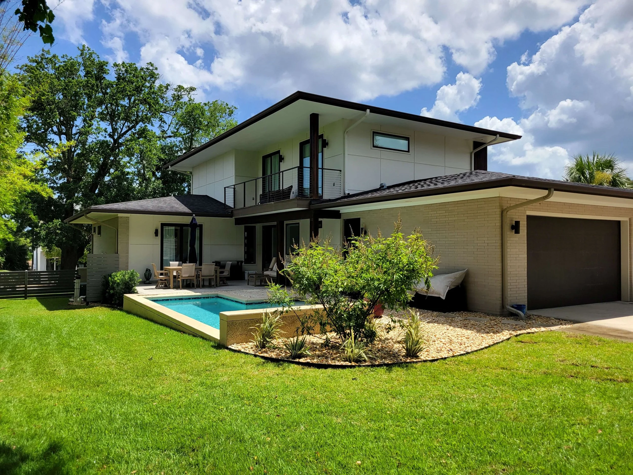 Modern two-story house with white exterior, rooftop balcony, backyard swimming pool, patio furniture, and landscaped lawn with small bushes and trees under partly cloudy sky.