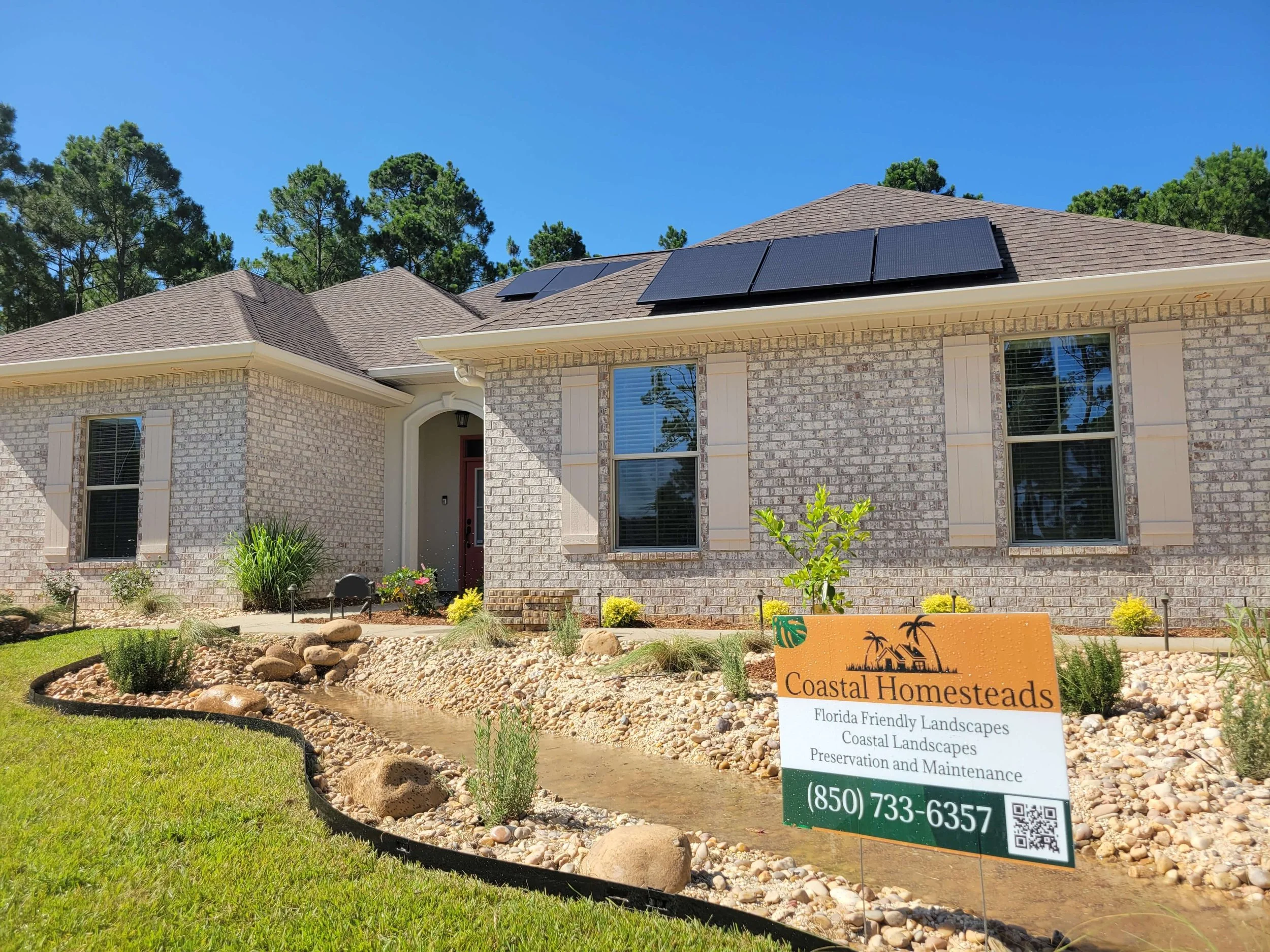 A single-story house with light-colored brick exterior, dark shingles, and solar panels on the roof. Front yard with landscaped rock garden, small water feature, and a sign for Coastal Homesteads, advertising Florida-friendly landscapes, coastal land