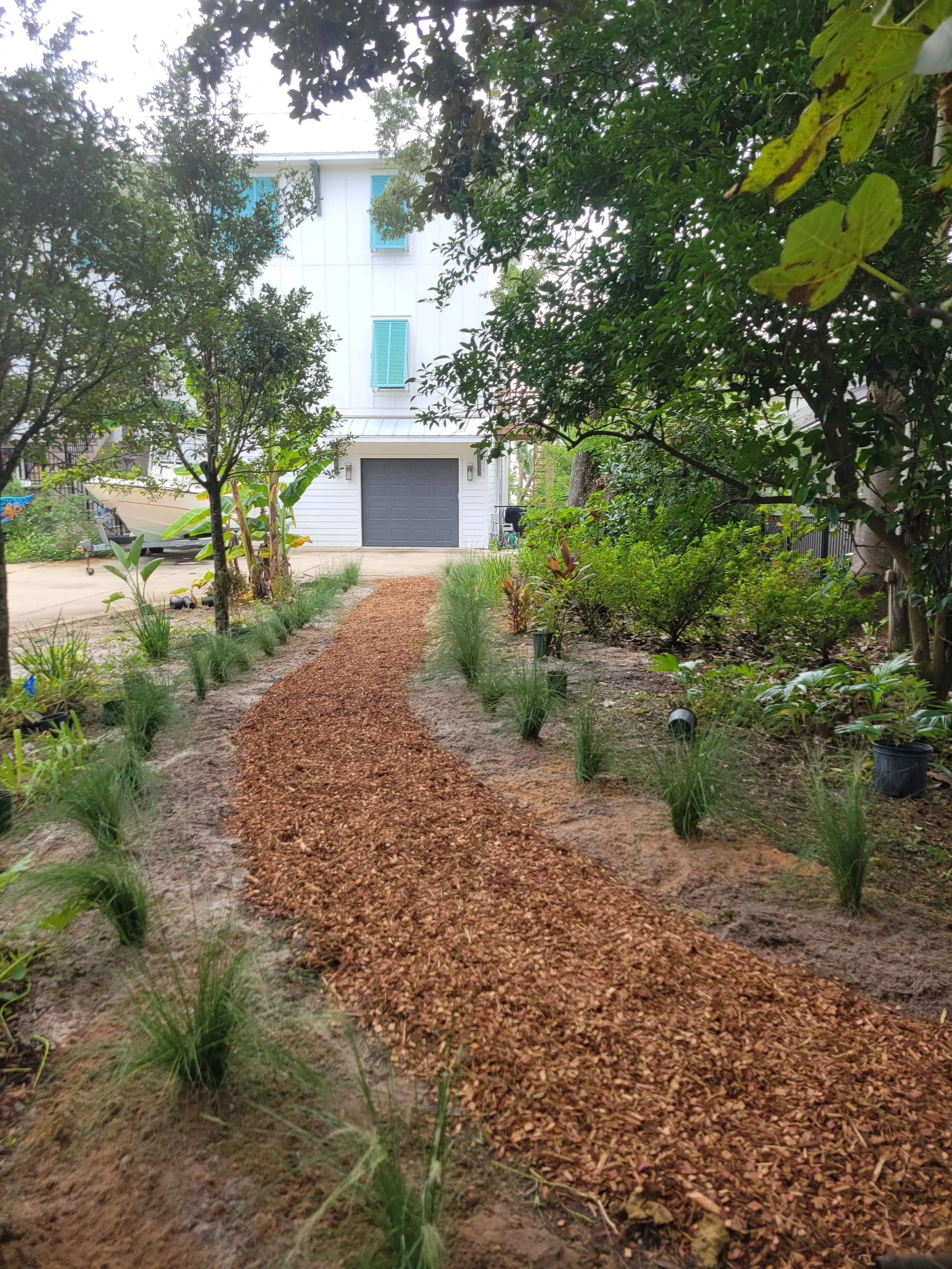 A landscaped backyard with a winding gravel path leading to a house with a garage. The yard has small plants, shrubs, and trees on both sides of the path.