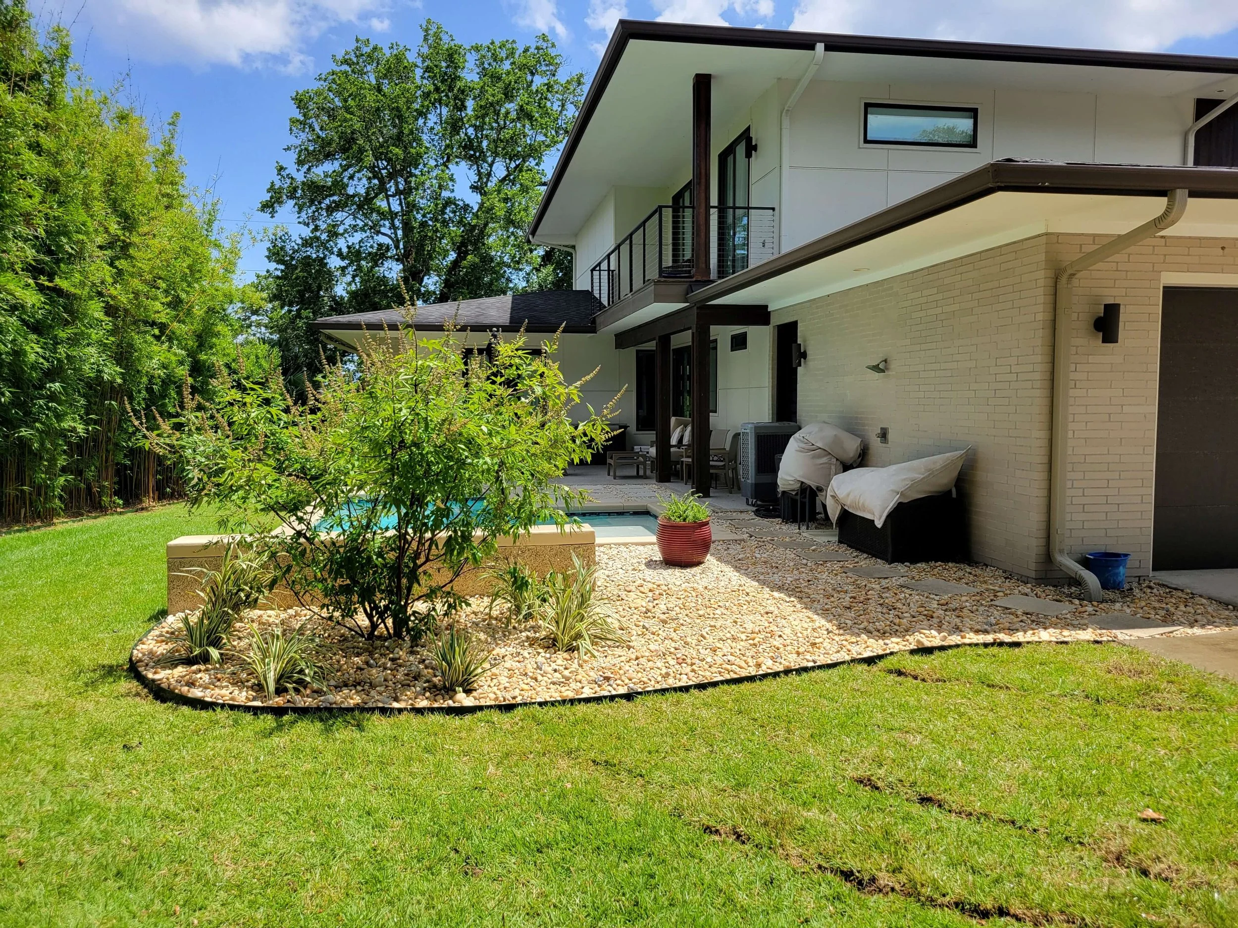 A backyard garden with a small tree, decorative plants, and gravel beside a modern house with an outdoor seating area and a swimming pool.