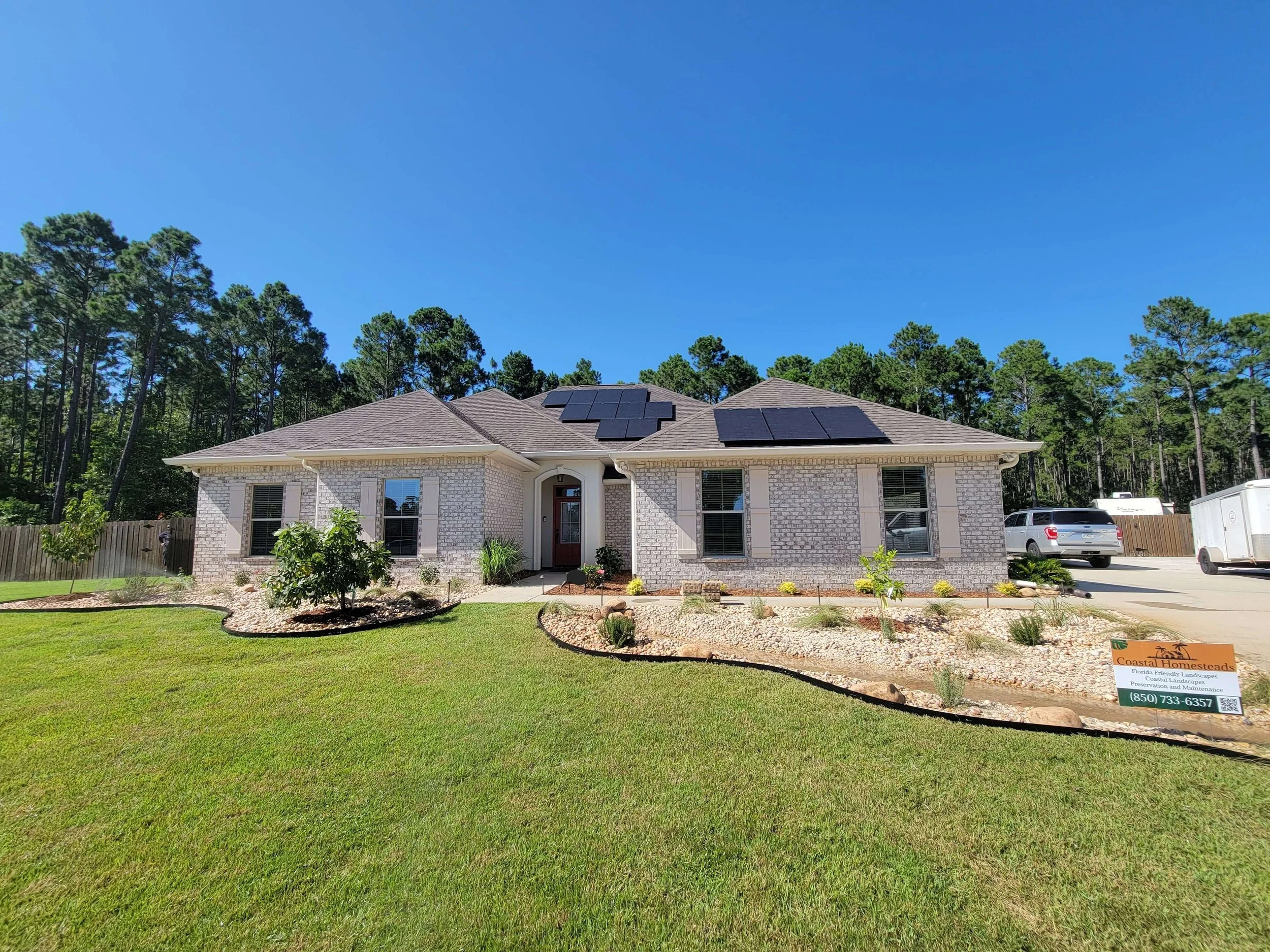 A newly built single-story house with solar panels on the roof, a well-maintained front yard with grass, trees, and shrubbery, and a driveway with parked vehicles and a sign in the yard advertising landscaping services.