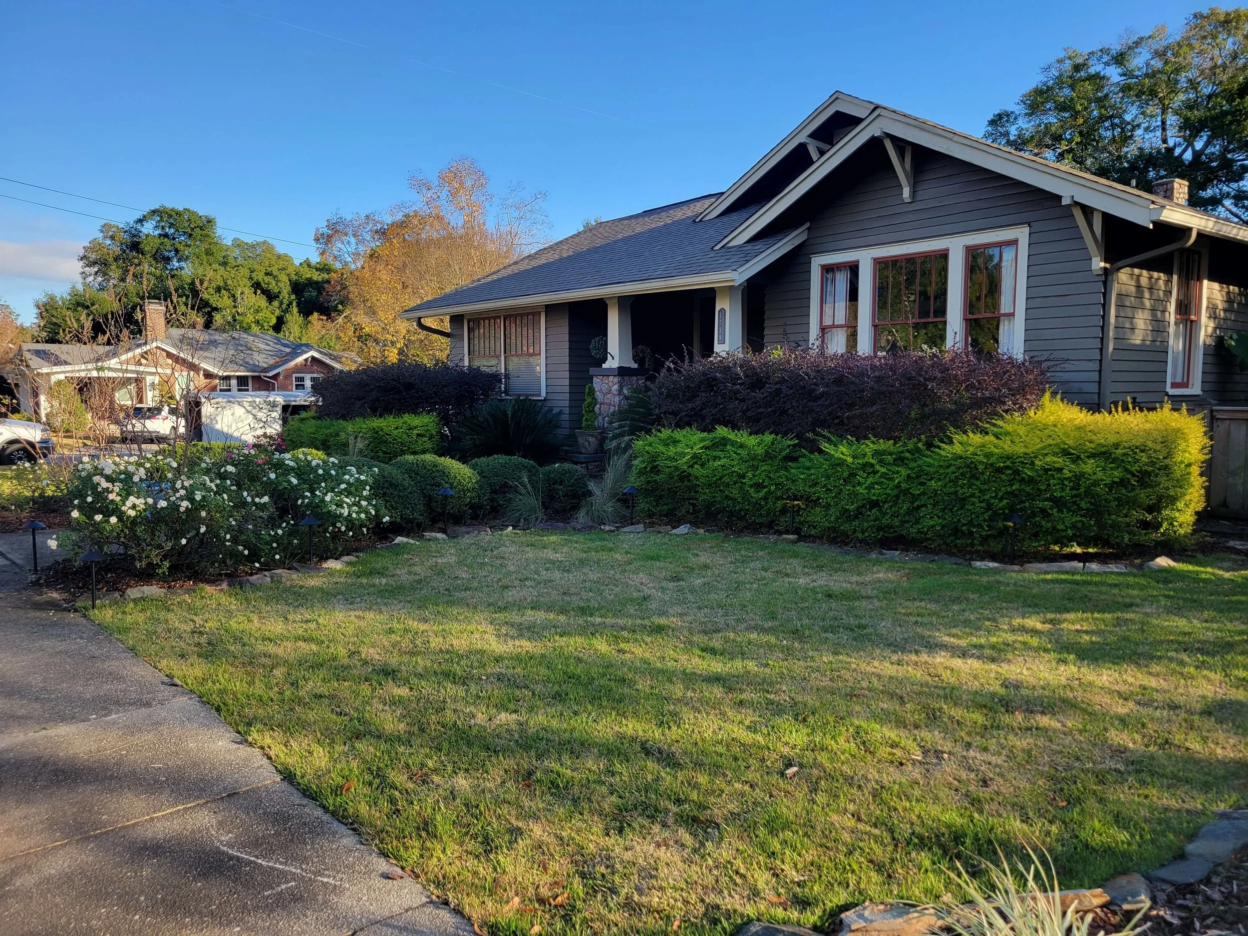 Front yard of a house with a well-maintained lawn, colorful flowering bushes, and shrubs under a clear blue sky.