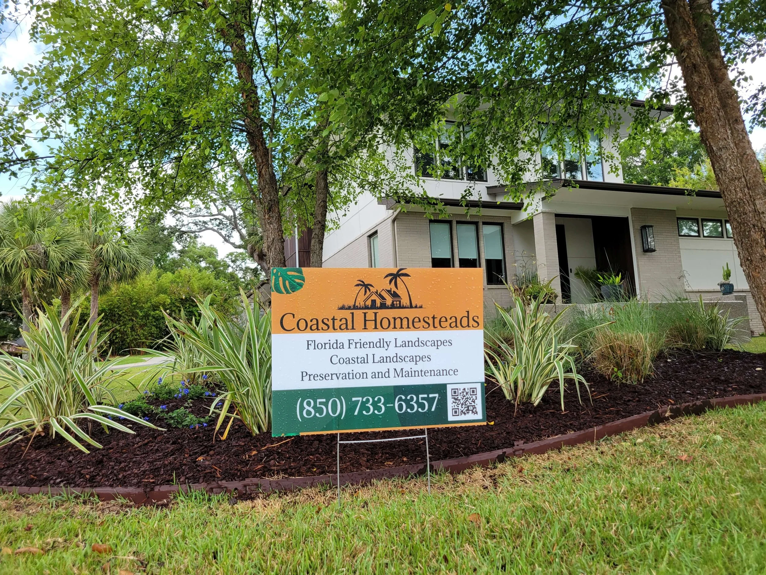 A sign in front of a modern house with trees and landscaped garden, displaying information about Coastal Homesteads and their services, including Florida-friendly landscapes, coastal landscapes, and preservation and maintenance, along with a phone nu
