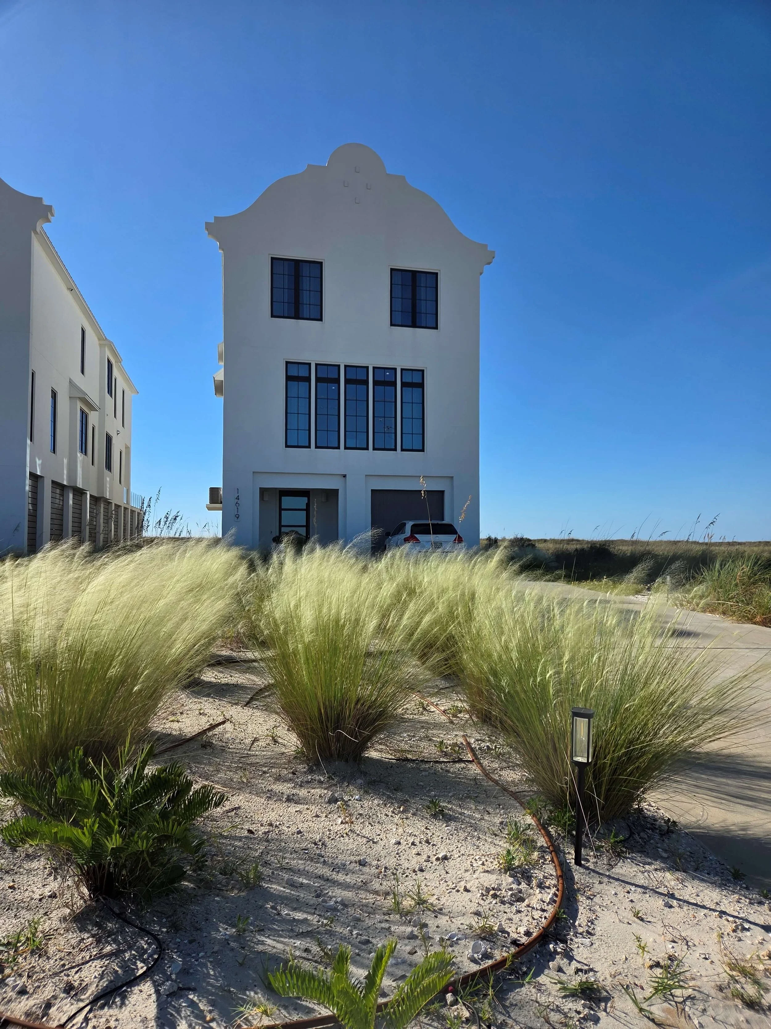 A white modern building with multiple windows, parked car, and sandy landscape with coastal grasses and a pathway under a clear blue sky.