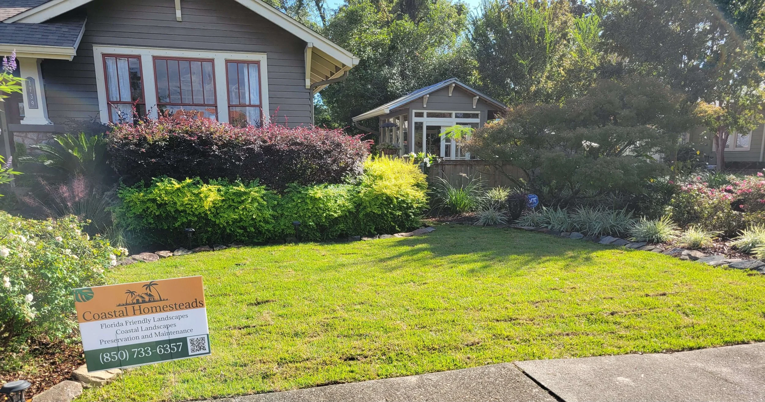 Front yard of a house with a well-maintained garden, various shrubs and plants, green lawn, sidewalk, and a sign for Coastal Homesteads indicating landscaping services.