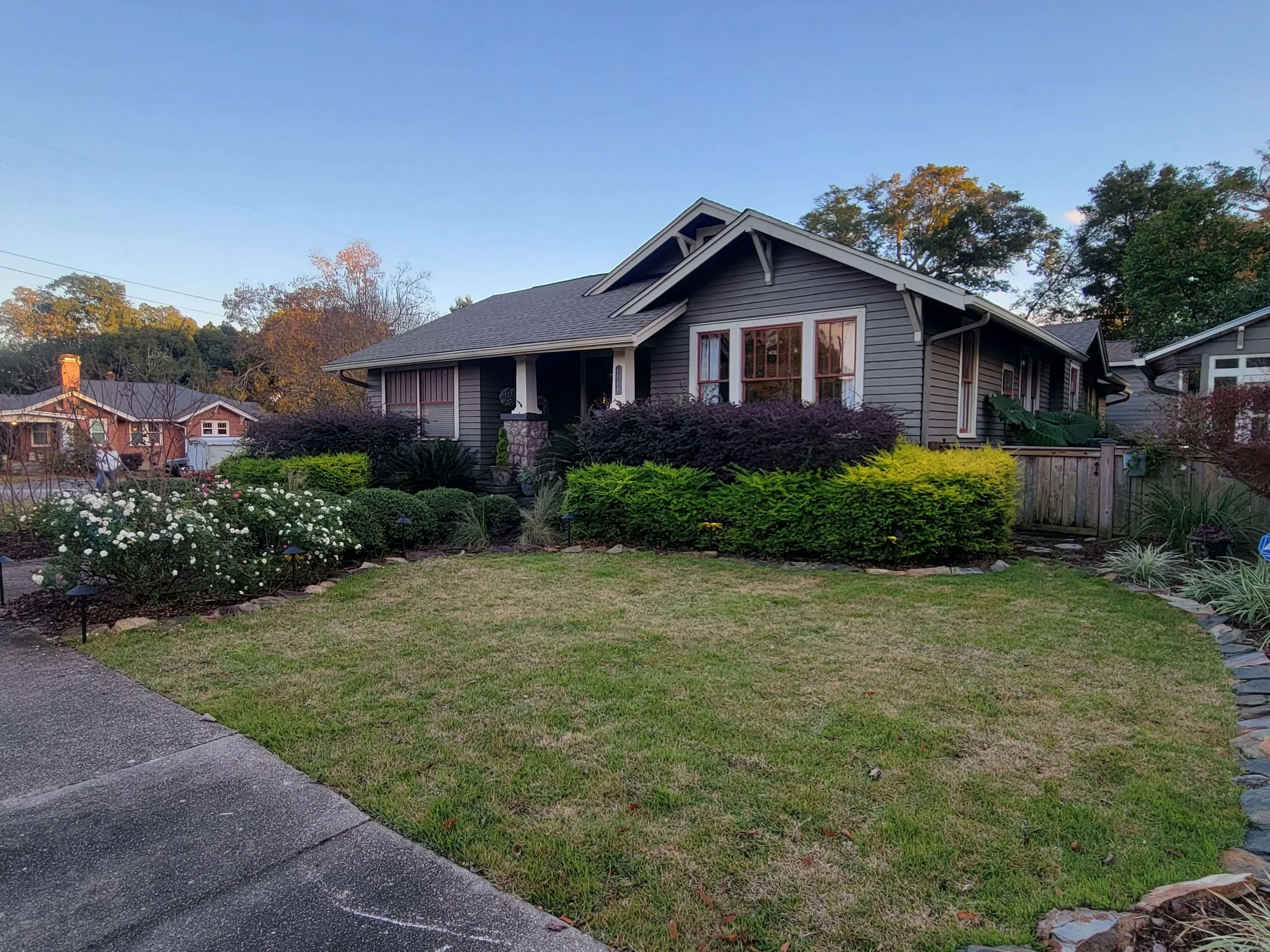 A house with a well-kept front yard, featuring lush shrubs, blooming flowers, and a mowed lawn, with neighboring houses and trees in the background during late afternoon.