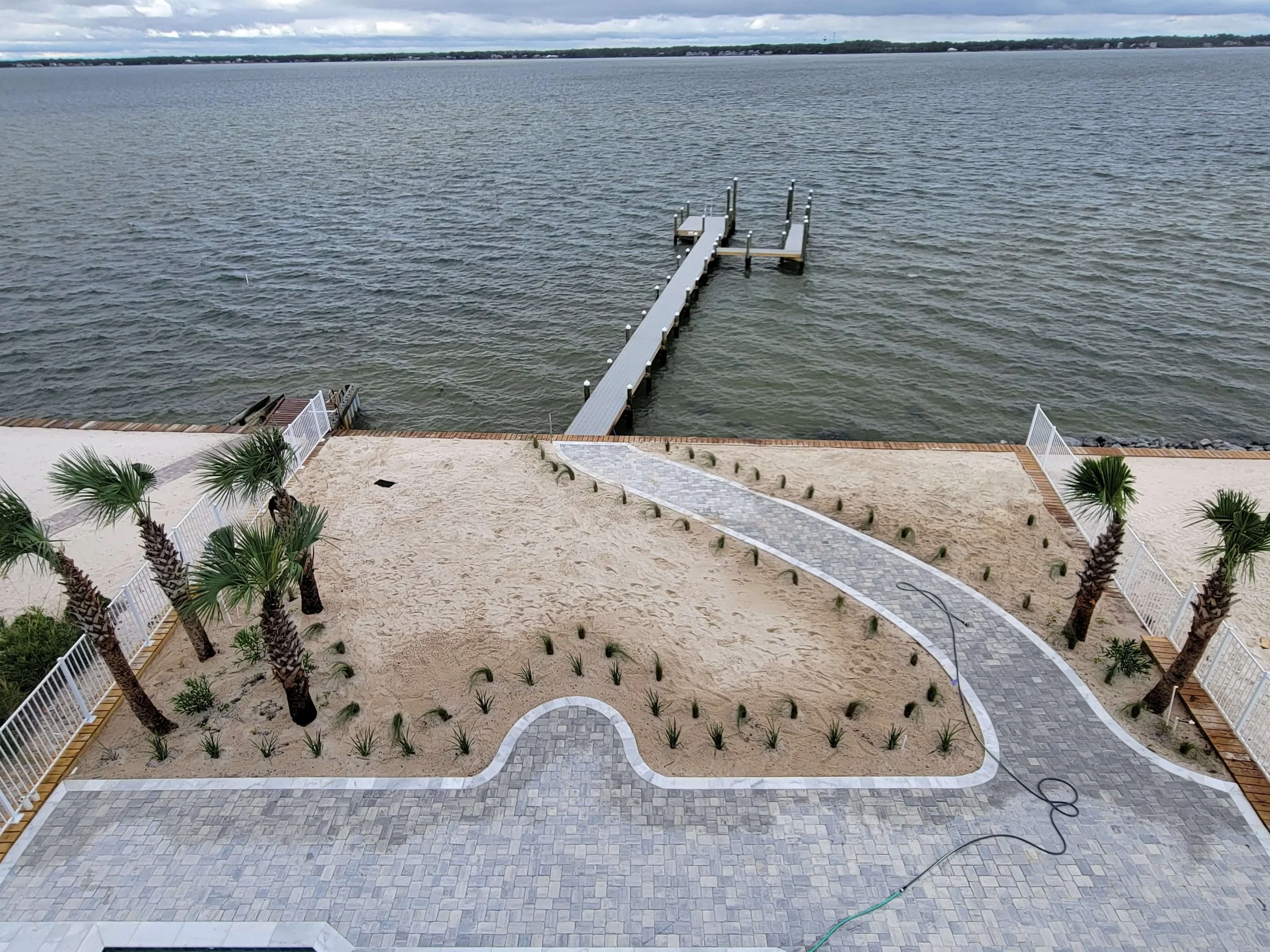 View of a backyard with palm trees, a paved walkway and a sandy area leading to a dock on a large body of water with a distant shoreline.