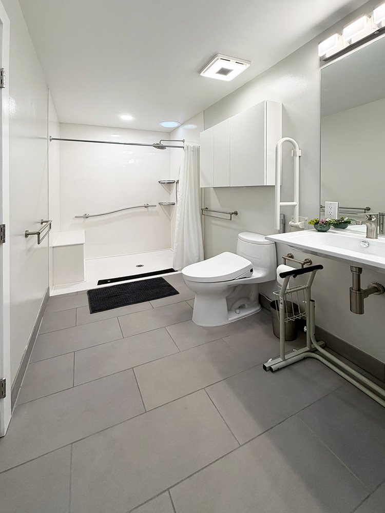 The primary suite bathroom. There is a roll-in shower, wall-mounted sink, toilet with a bidet seat and various grab bars. 