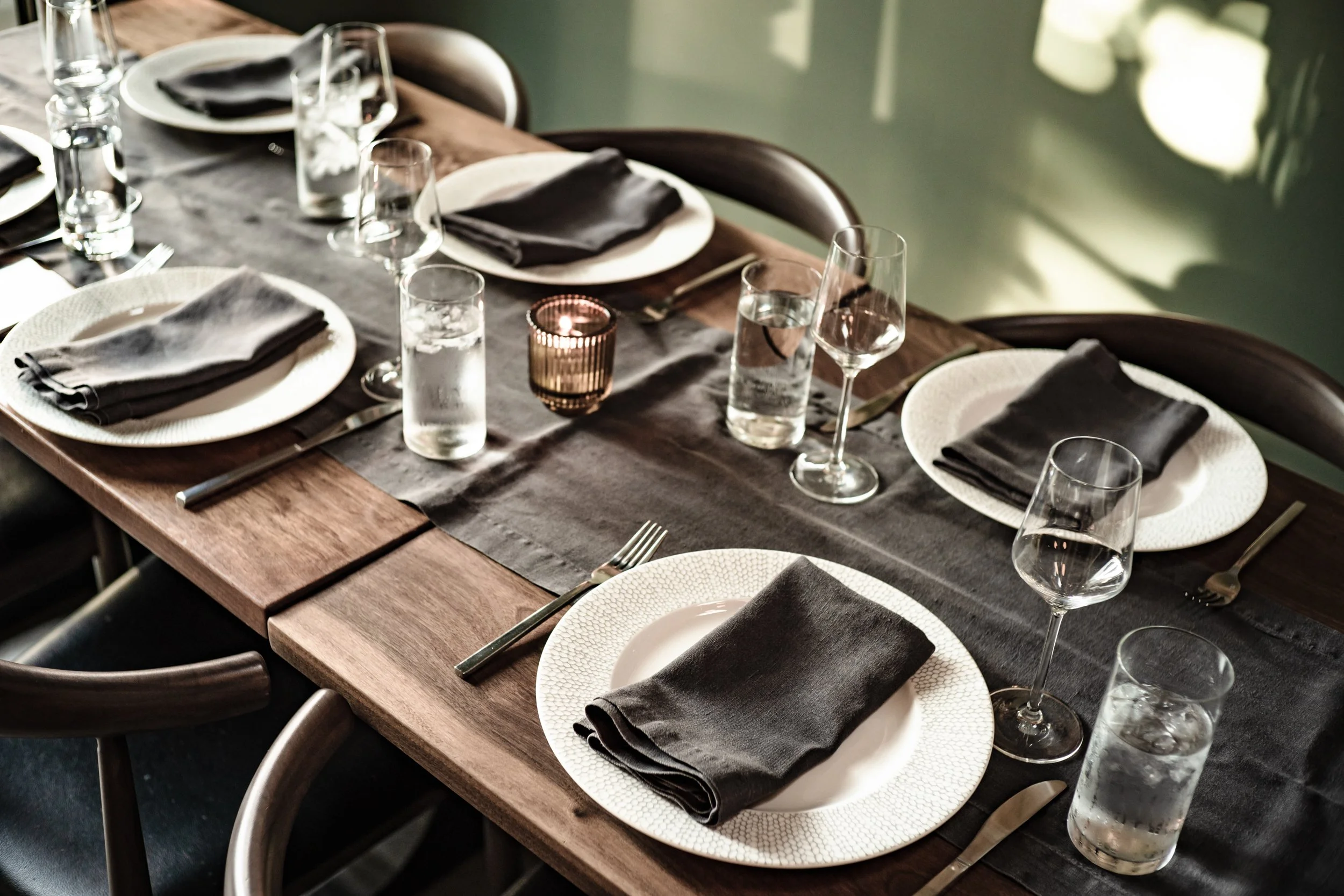 A dining table set for six with white plates, black cloth napkins, wine glasses, water glasses, and a candle in a decorative holder in the center.
