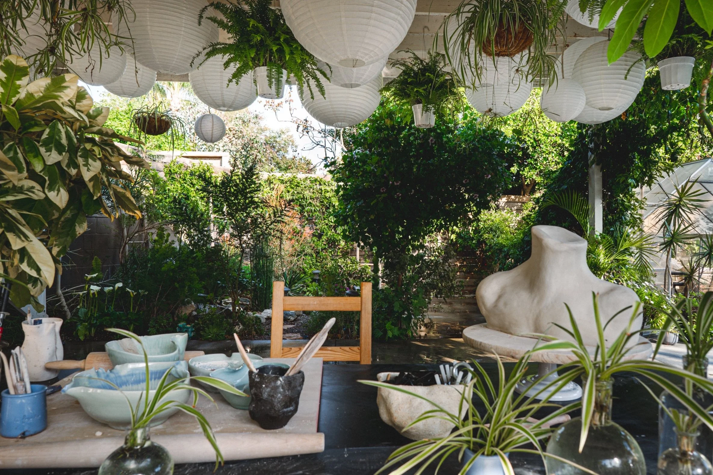 A lush outdoor garden workspace decorated with hanging paper lanterns, surrounded by green plants and trees, with a large abstract sculpture and a table set with ceramic bowls, utensils, and potted plants.