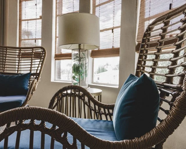 Interior of a cozy room with wicker chairs, blue cushions, a glass lamp with a green plant, and windows with bamboo blinds.