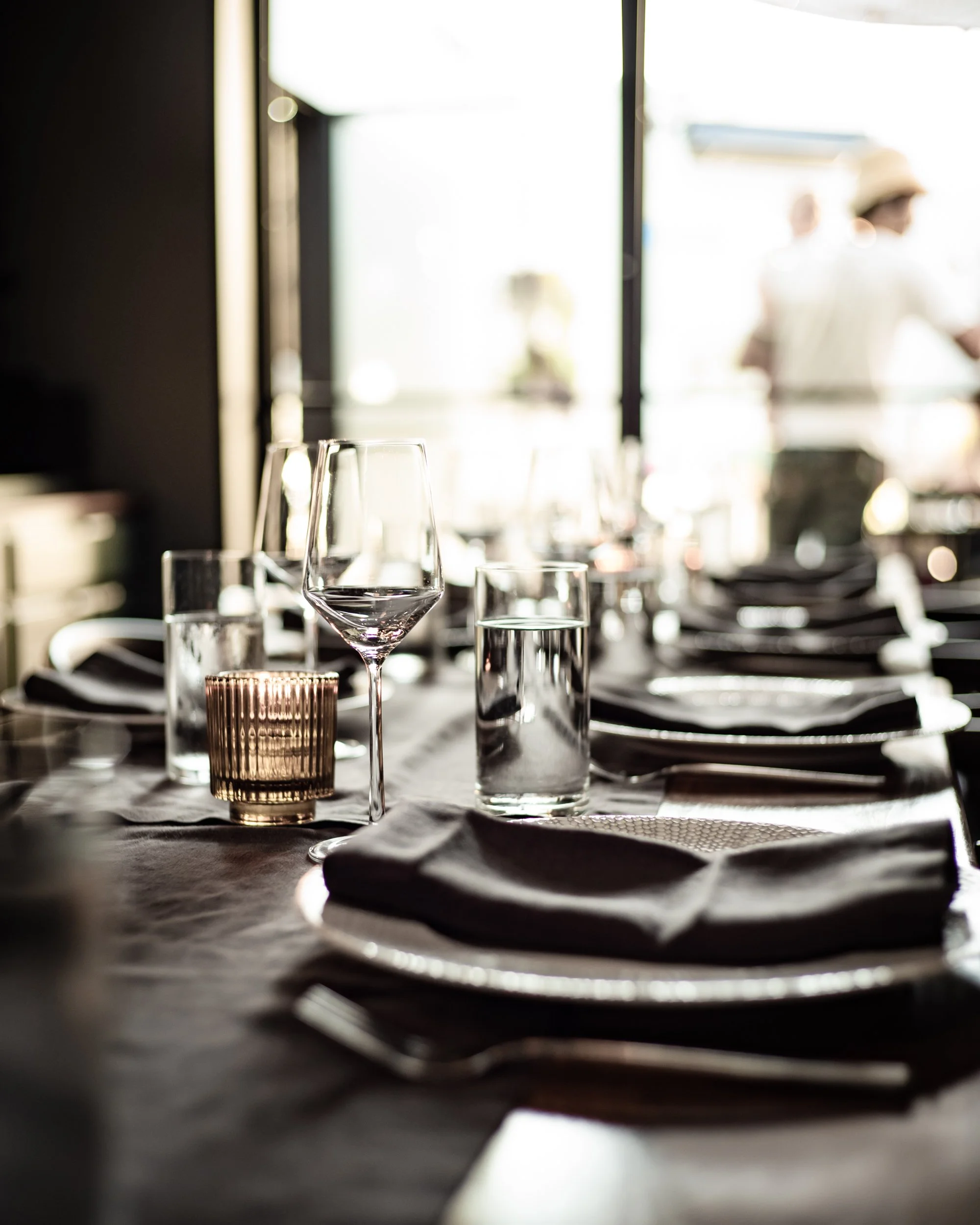 A close-up of a elegant dining table set with wine glasses, water glasses, silverware, and napkins in a well-lit restaurant with large windows.