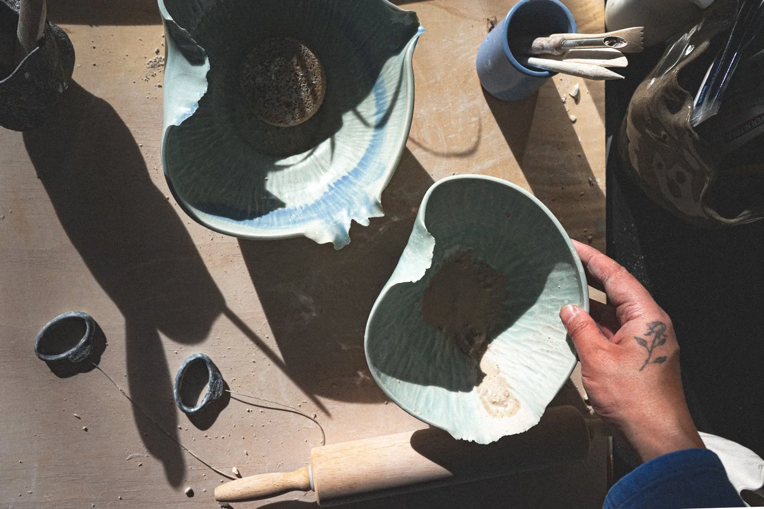 A person with a tattoo on their hand holds a broken ceramic bowl over a work surface with other broken bowls, a rolling pin, and tools in a pottery studio.