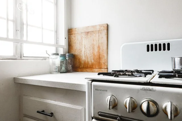 A kitchen corner with a white gas stove, a wooden cutting board, and glass jars on the counter beside a window with natural light.