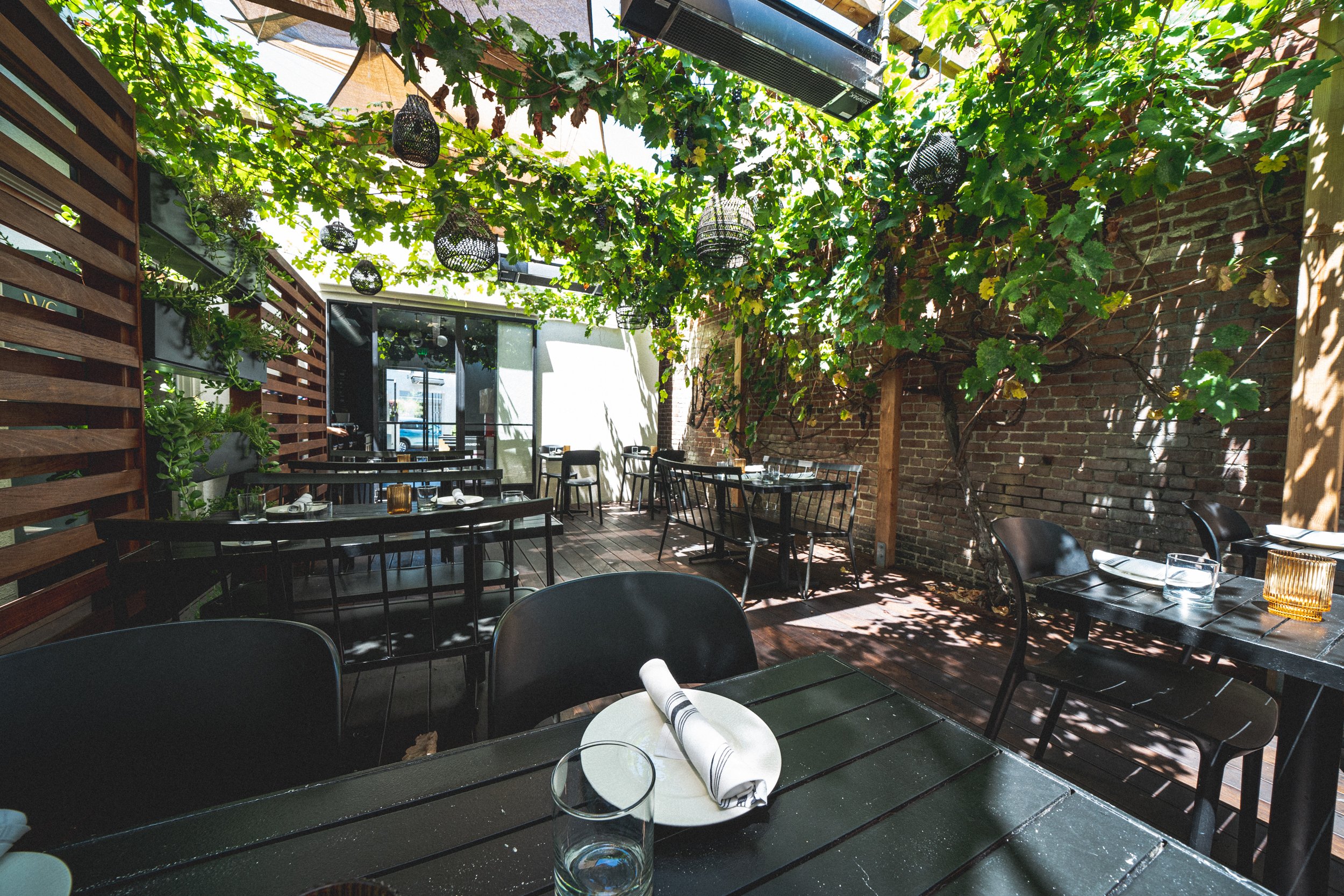Outdoor restaurant patio with black tables and chairs, surrounded by greenery and a brick wall, with sunlight filtering through leaves and hanging lanterns.