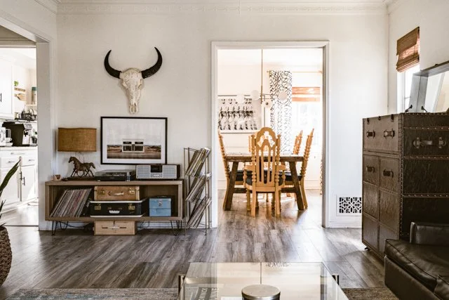 Living room with a wall-mounted bull skull, a sideboard with framed art and decorative items, a wooden dining table with chairs, and a doorway leading to the kitchen.