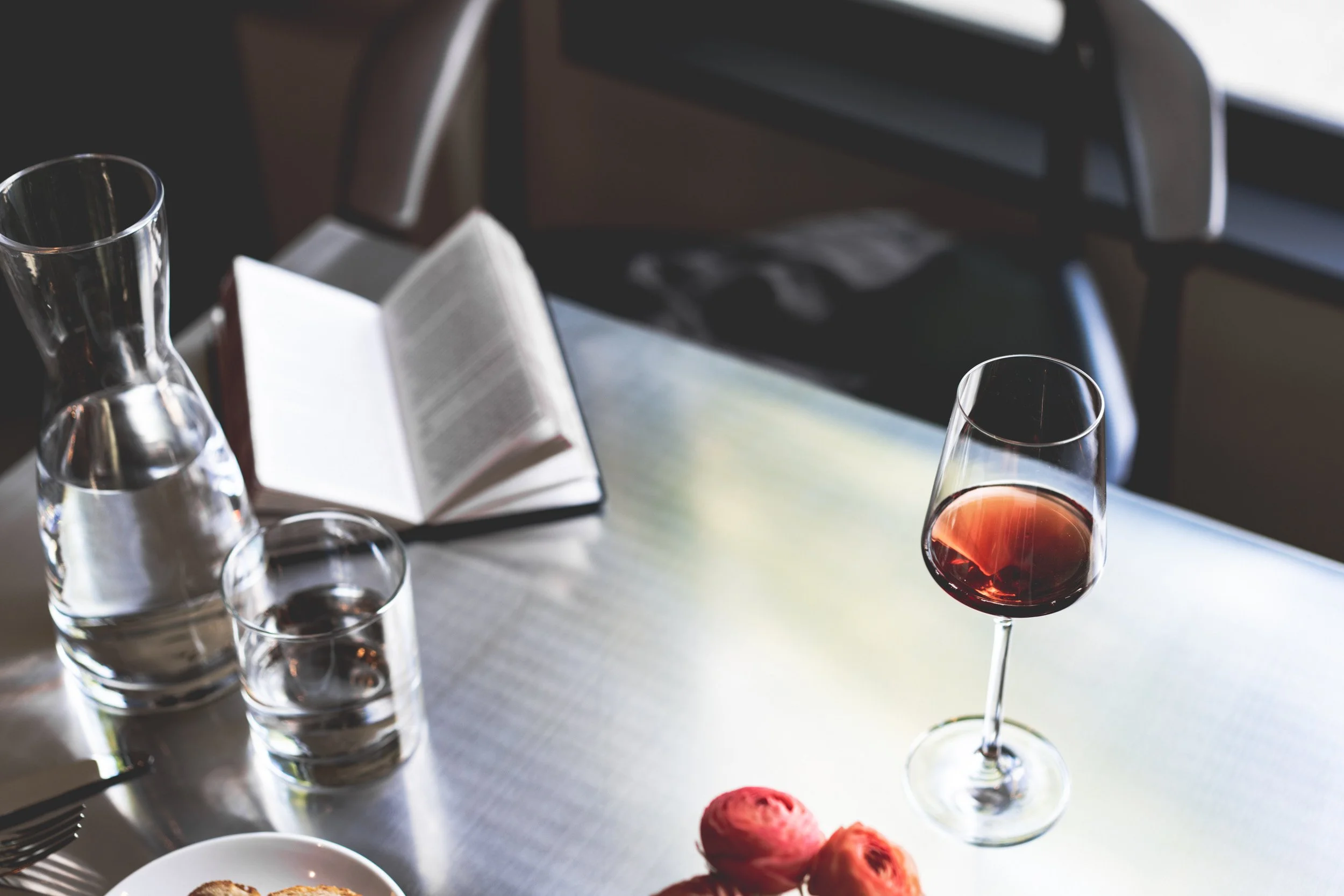 A dining table with a glass of red wine, a glass pitcher filled with water, two glasses of water, an open book, a plate of food, and some small desserts on the table.