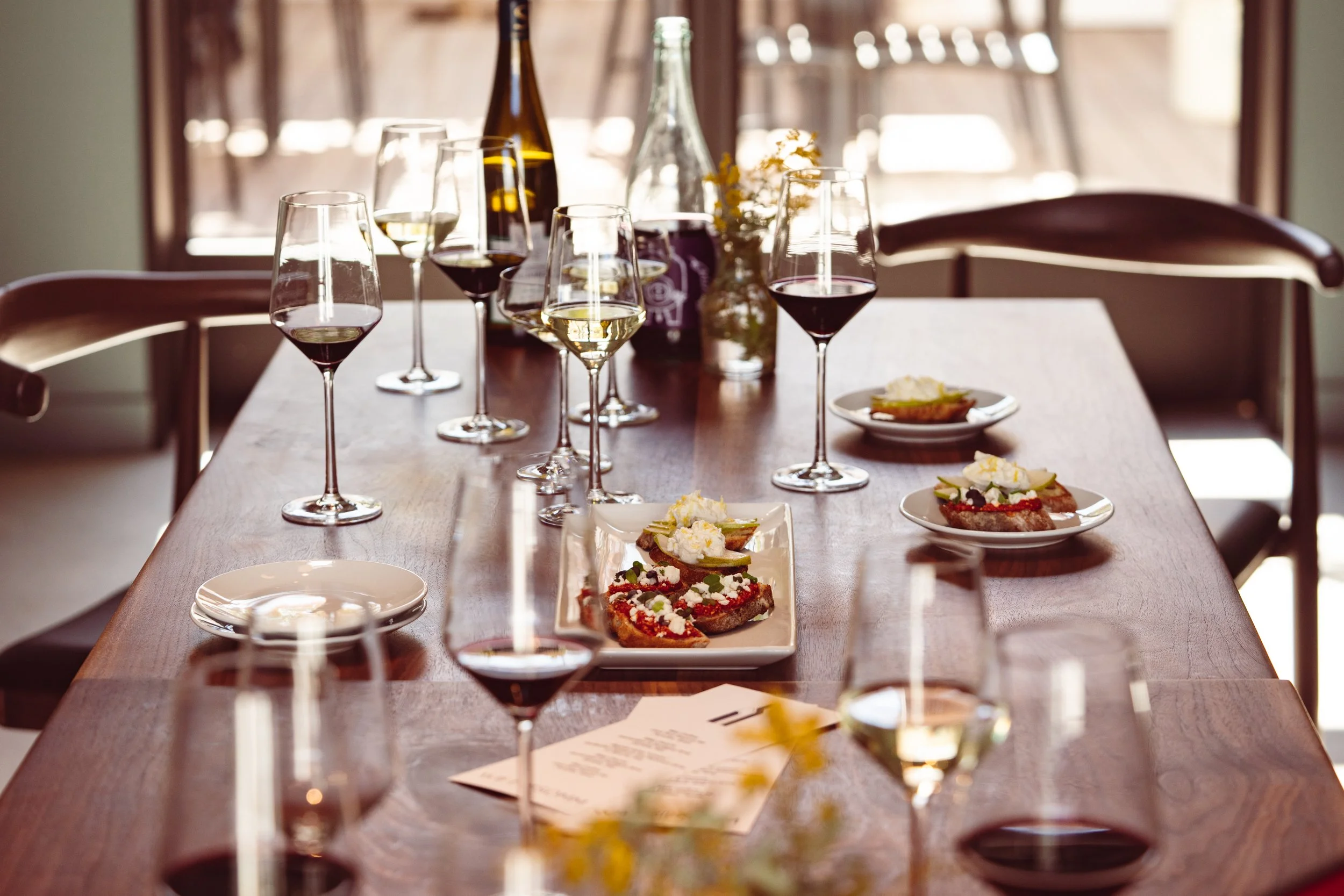 A dining table set for a wine tasting or small gathering, with several glasses of red and white wine, plates of appetizers, and a menu or note on the table in a sunlit room.
