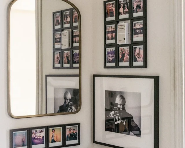 A wall decorated with multiple framed photographs, including a mirror and black-and-white images of a person with a photo prop, arranged in a collage style.