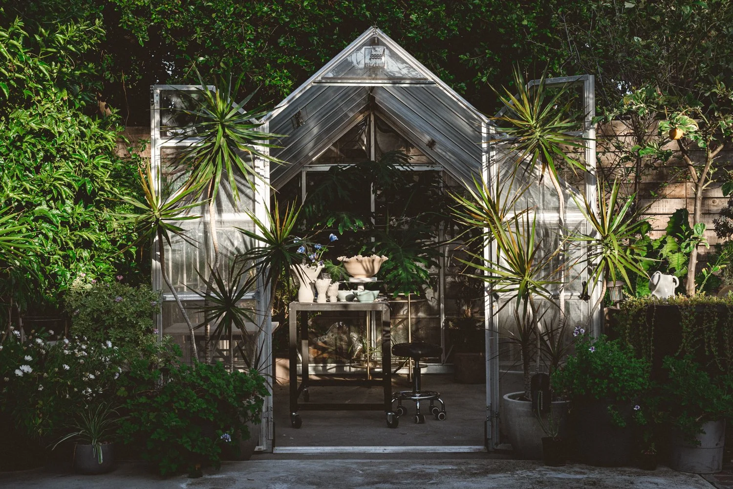 A small greenhouse made of metal and glass surrounded by potted plants and trees outside.