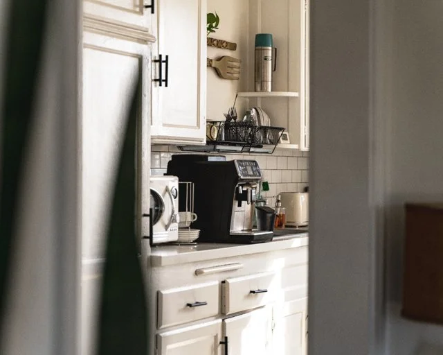 Partial view of a kitchen with white cabinets, a coffee maker, toaster, and some kitchen utensils on a countertop. A small potted plant and a thermos are on the shelves.
