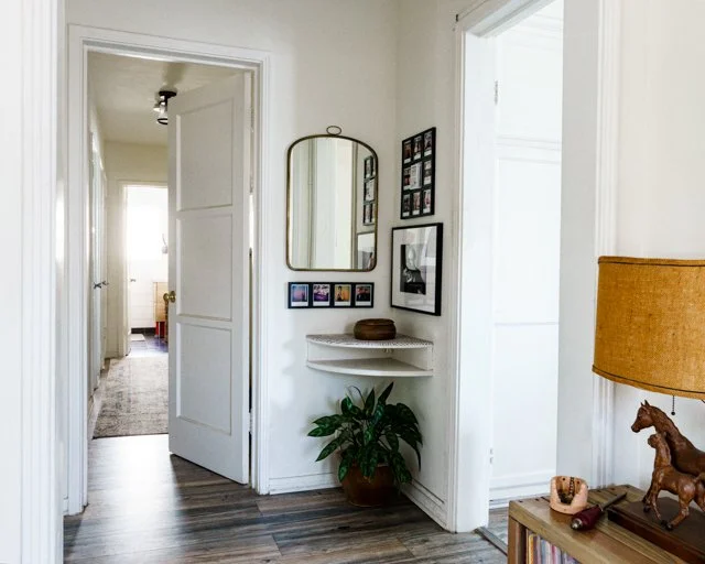 Interior view of a house hallway with a white door open to a room and a curved semi-circle white shelf below wall decor and a potted plant on the floor.